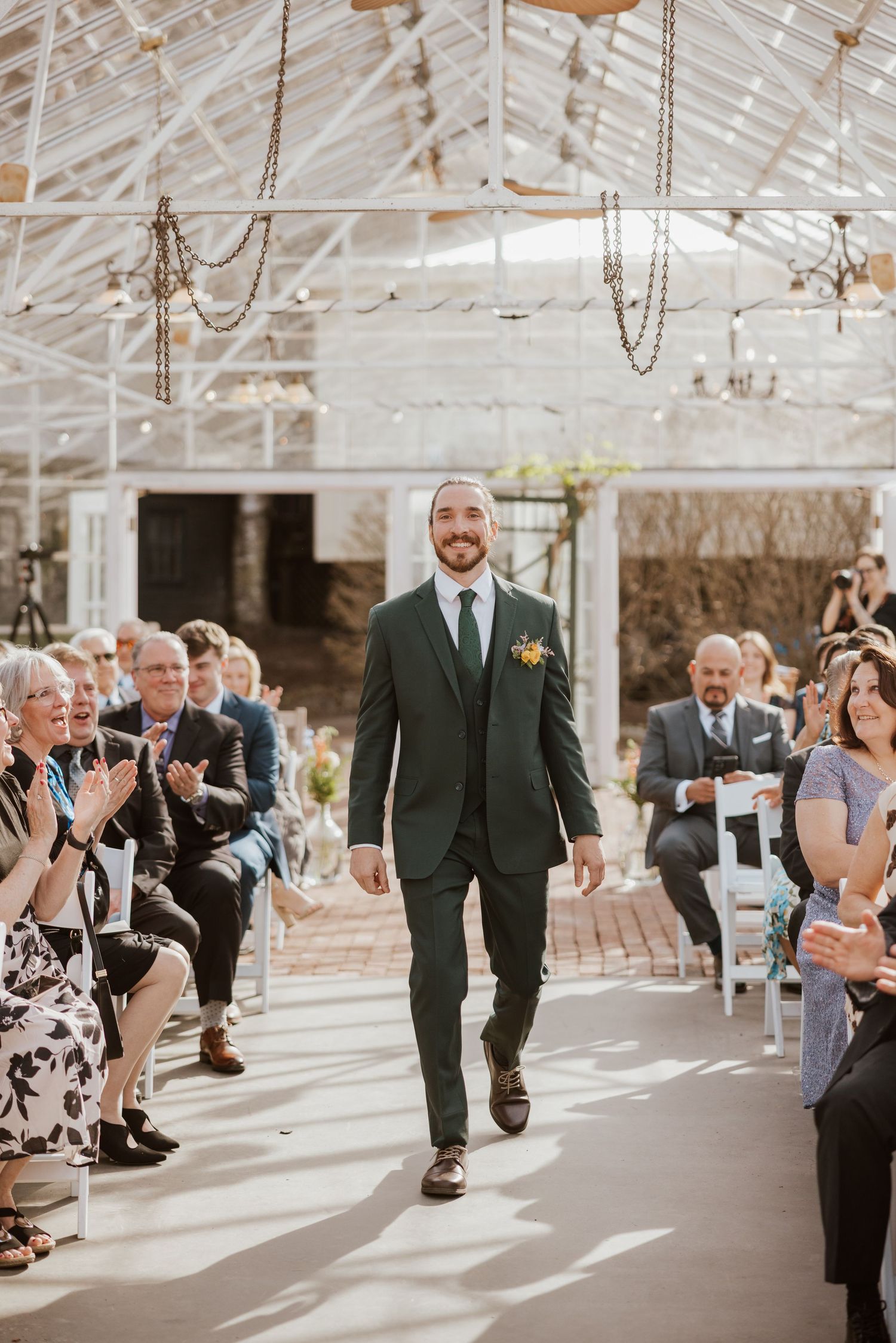 Smiling groom in dark green suit walks down wedding aisle in glass greenhouse venue with seated guests applauding.