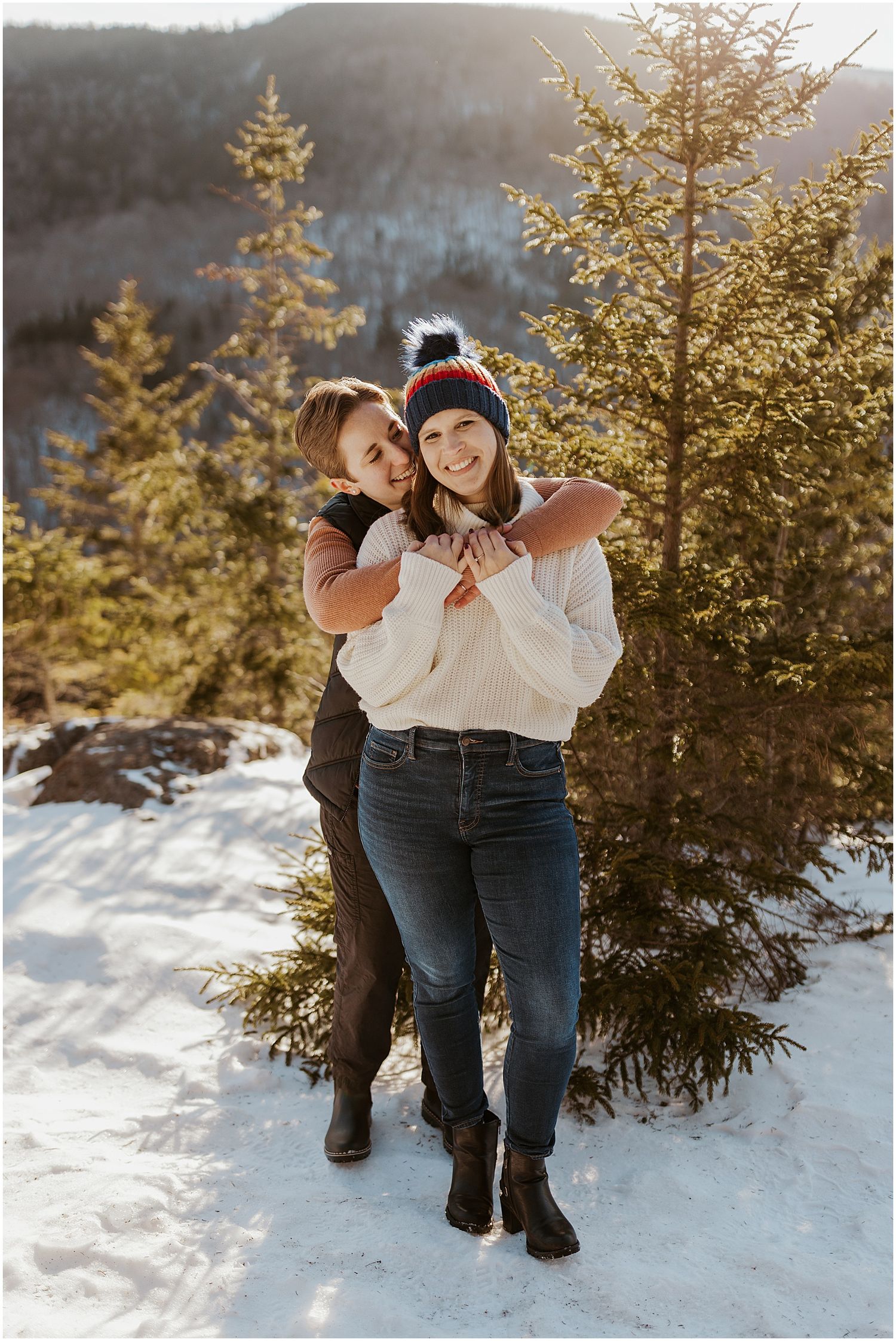 A playful winter engagement photo in the snow with evergreen trees and mountain views.