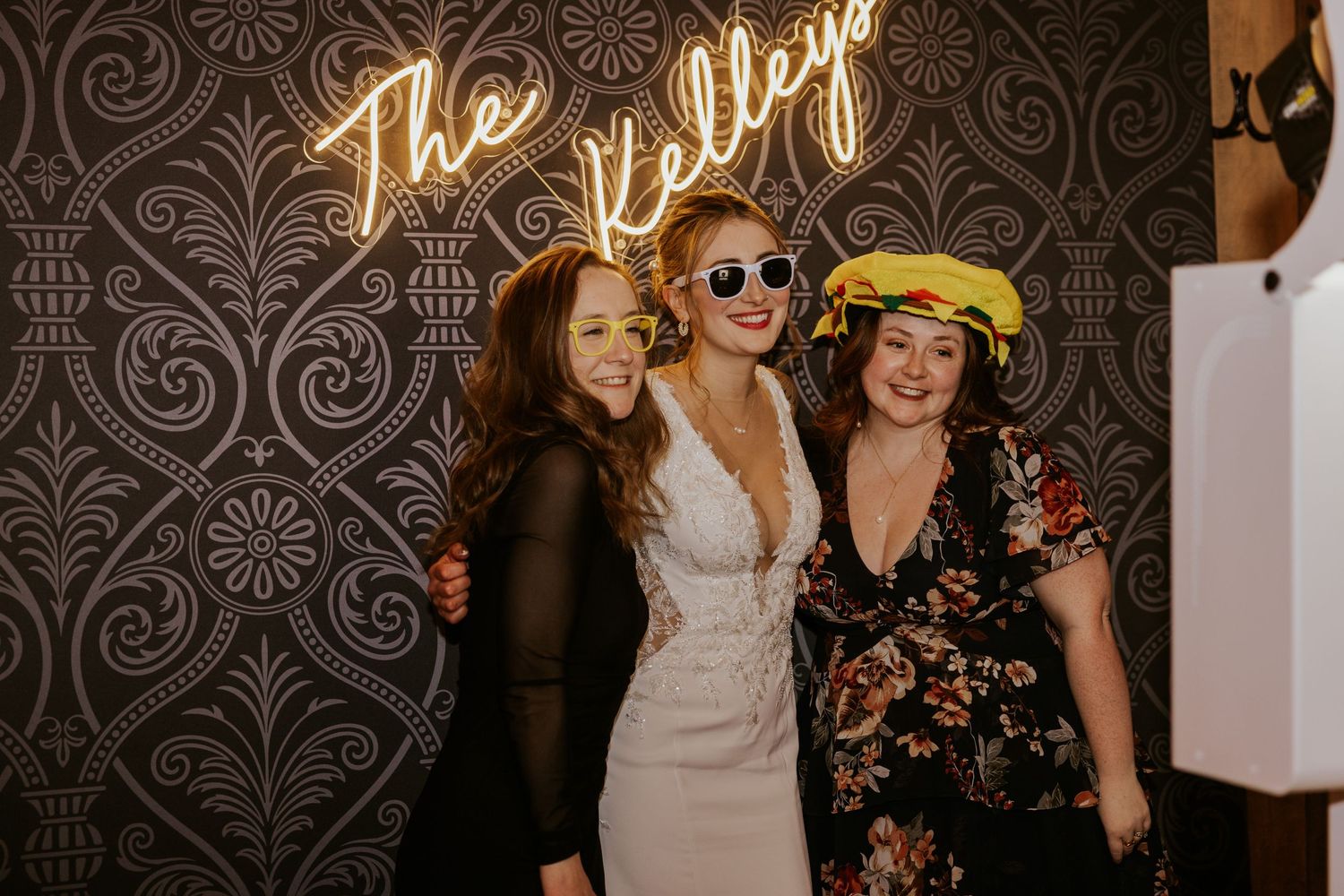 Three women posing and smiling in front of a decorative wall with a neon sign reading The Kelleys.
