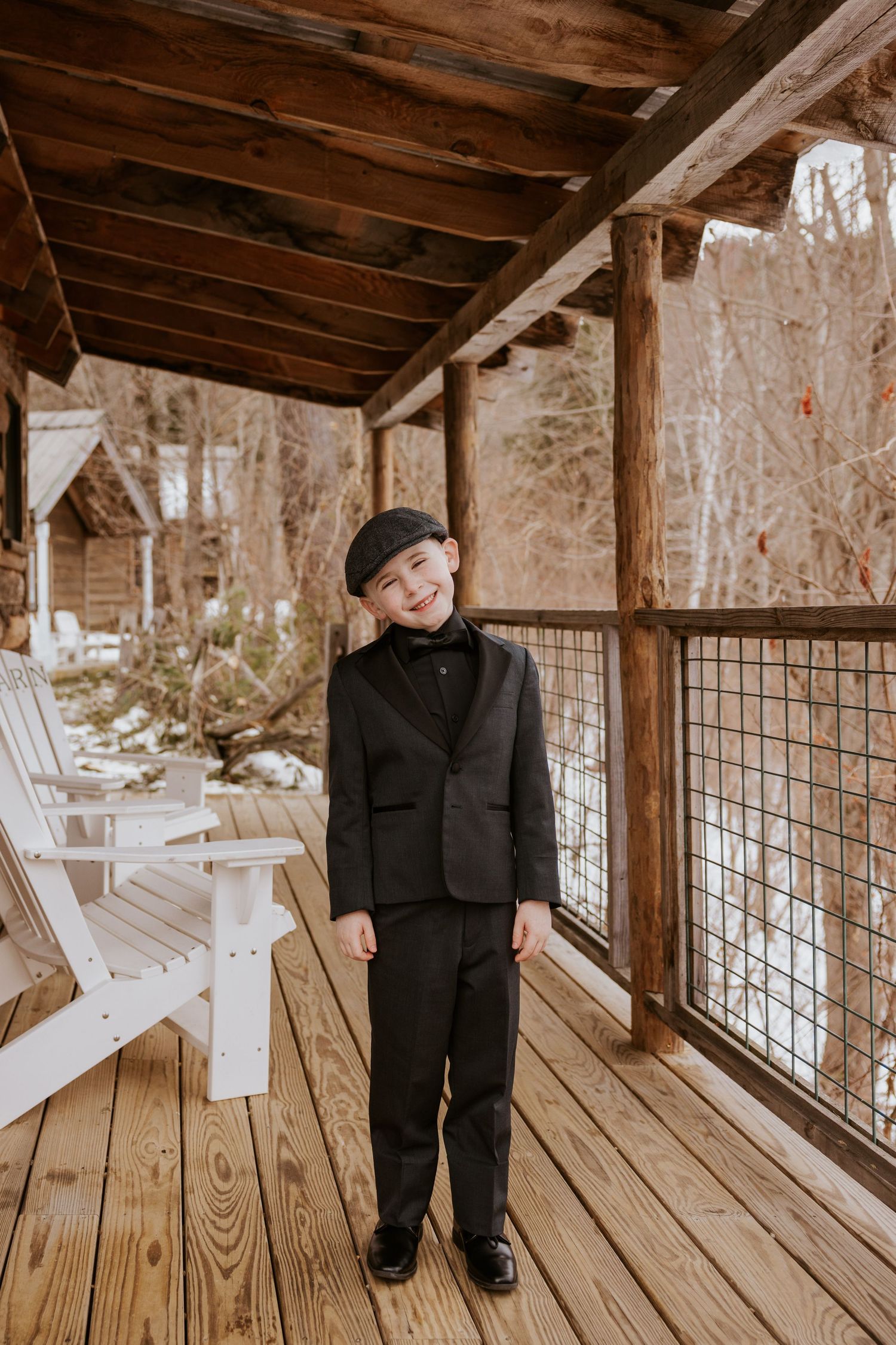 Young ring bearer in black suit and cap smiling on rustic wooden porch in winter.