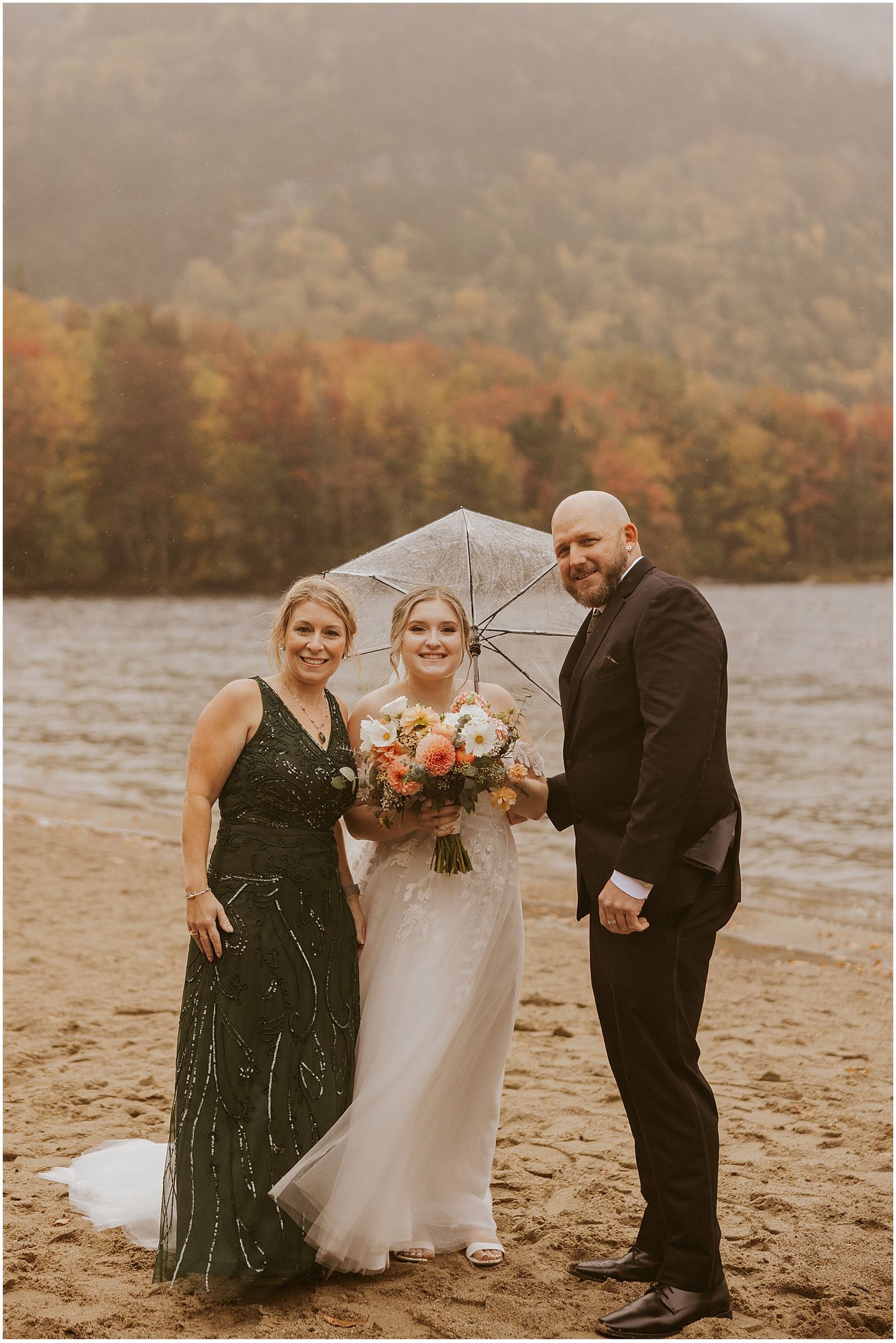 Three wedding attendees pose on a beach with umbrella against fall foliage and mountain backdrop.