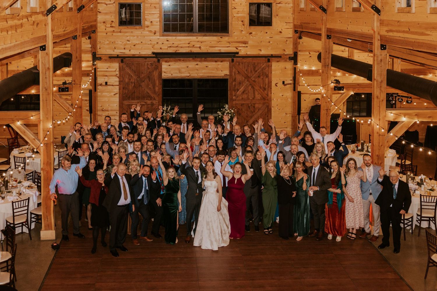 Wedding guests dance and celebrate in a rustic wooden barn venue illuminated by warm string lights.