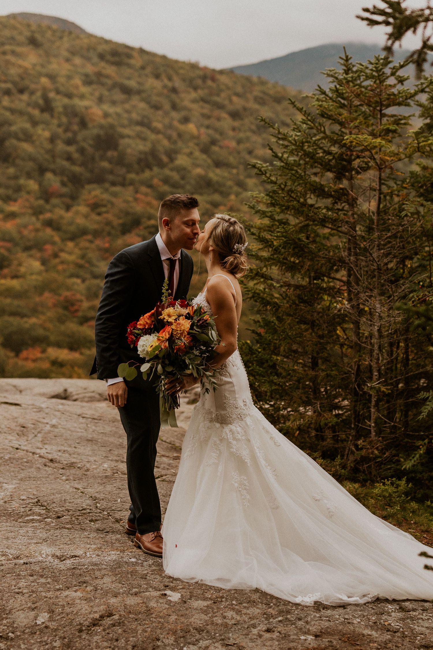 A couple shares an intimate moment on a mountain trail surrounded by evergreen trees and autumn foliage.