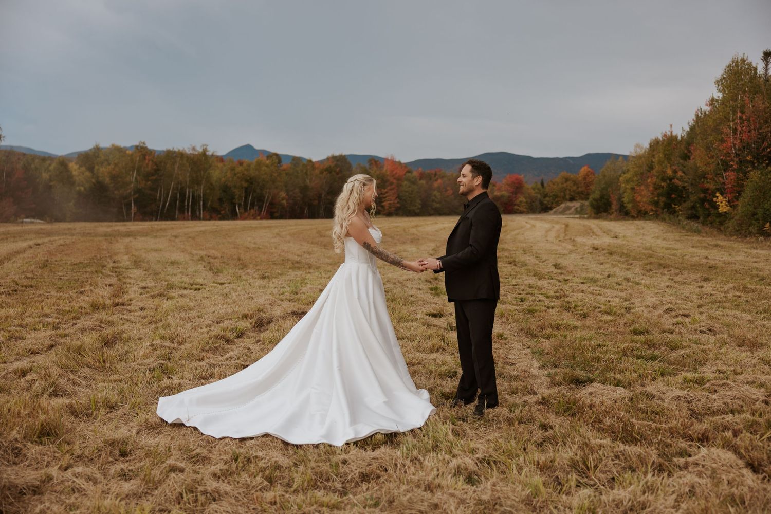 A bride and groom hold hands in a golden autumn field with colorful foliage and misty mountains in the background.