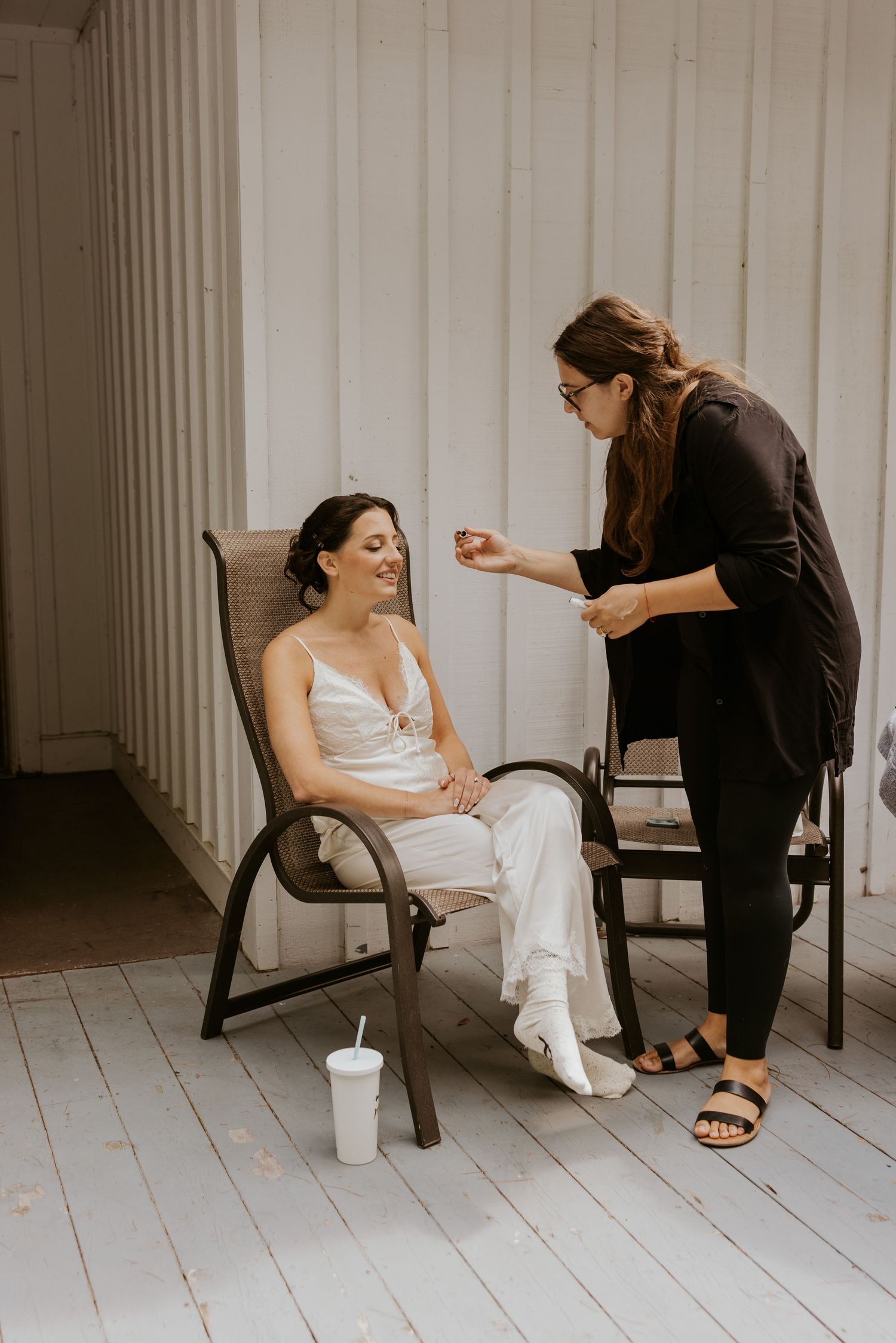 Bride in white slip dress smiles as makeup artist applies finishing touches on outdoor porch.