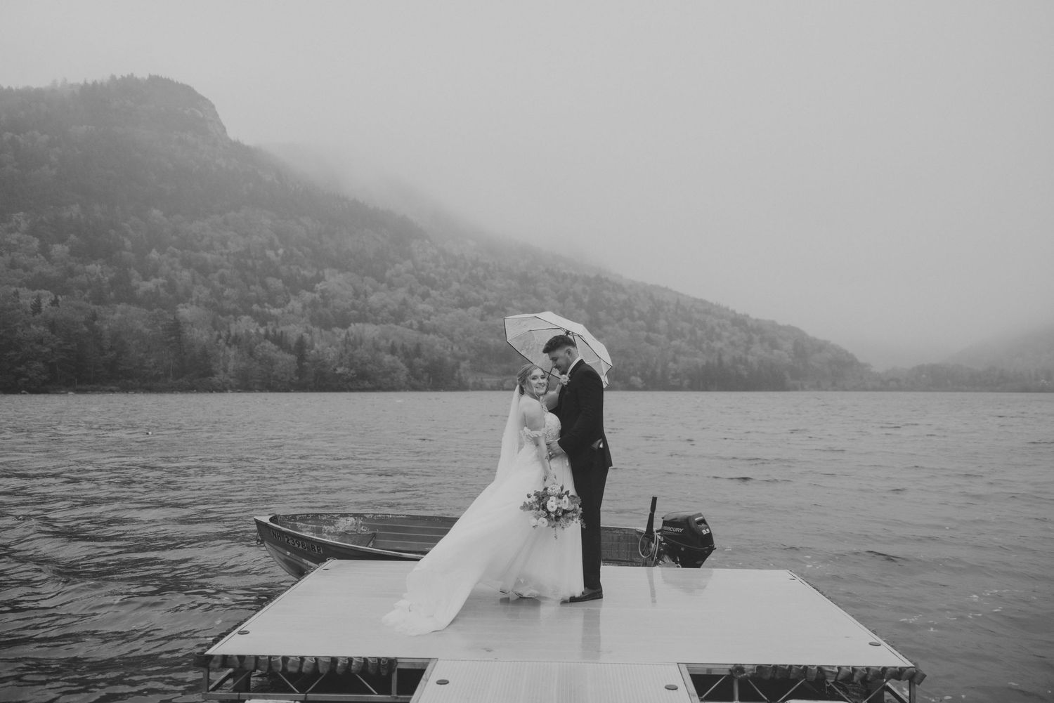 Black and white photo of bride and groom embracing on a misty lake dock with a rowboat nearby.