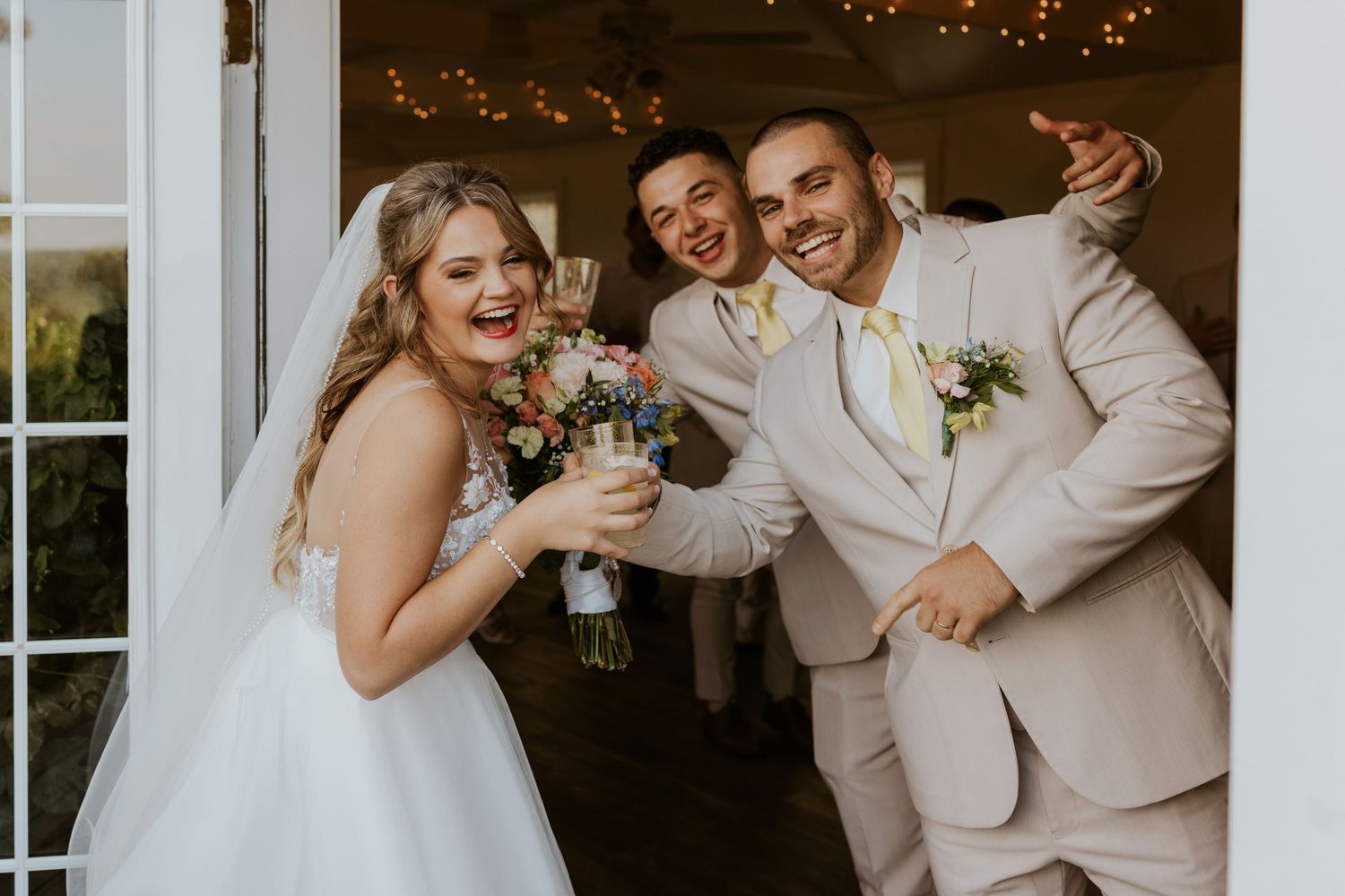 Joyful bride and groom celebrating with a guest at wedding venue entrance, bride holding colorful bouquet.