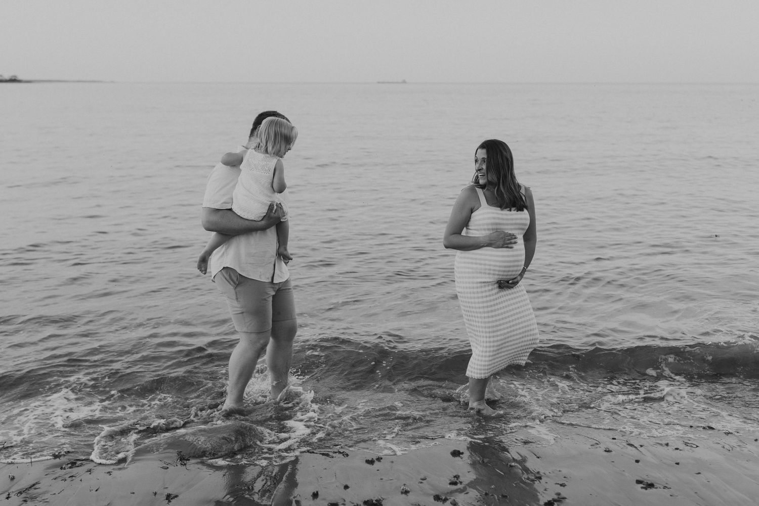 Black and white maternity photo of couple with toddler wading in ocean at sunset.