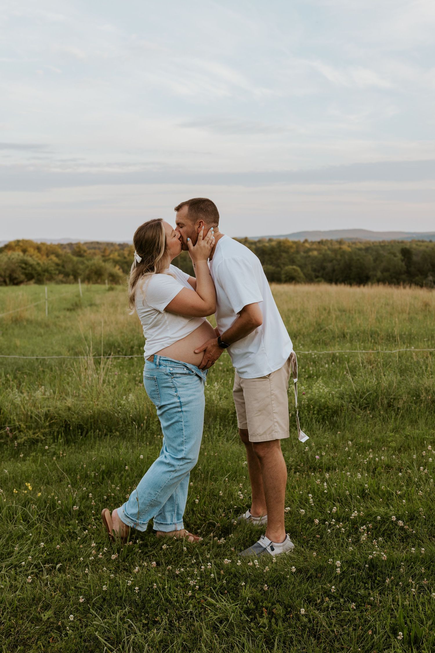 Pregnant couple sharing a kiss in an open grassy field, man gently touching woman's baby bump at golden hour.