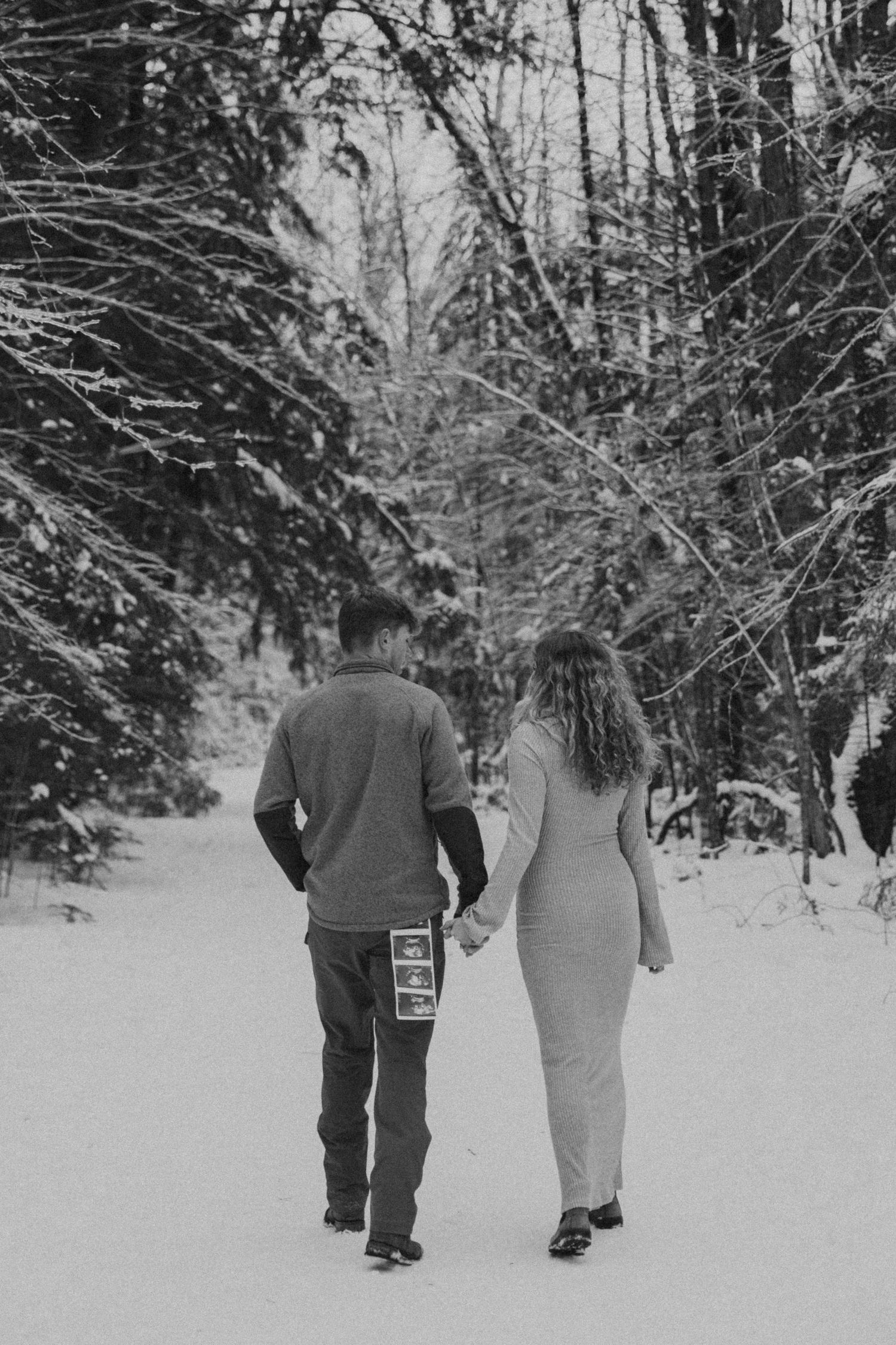 Black and white photo of expecting couple walking hand in hand through snowy forest path.