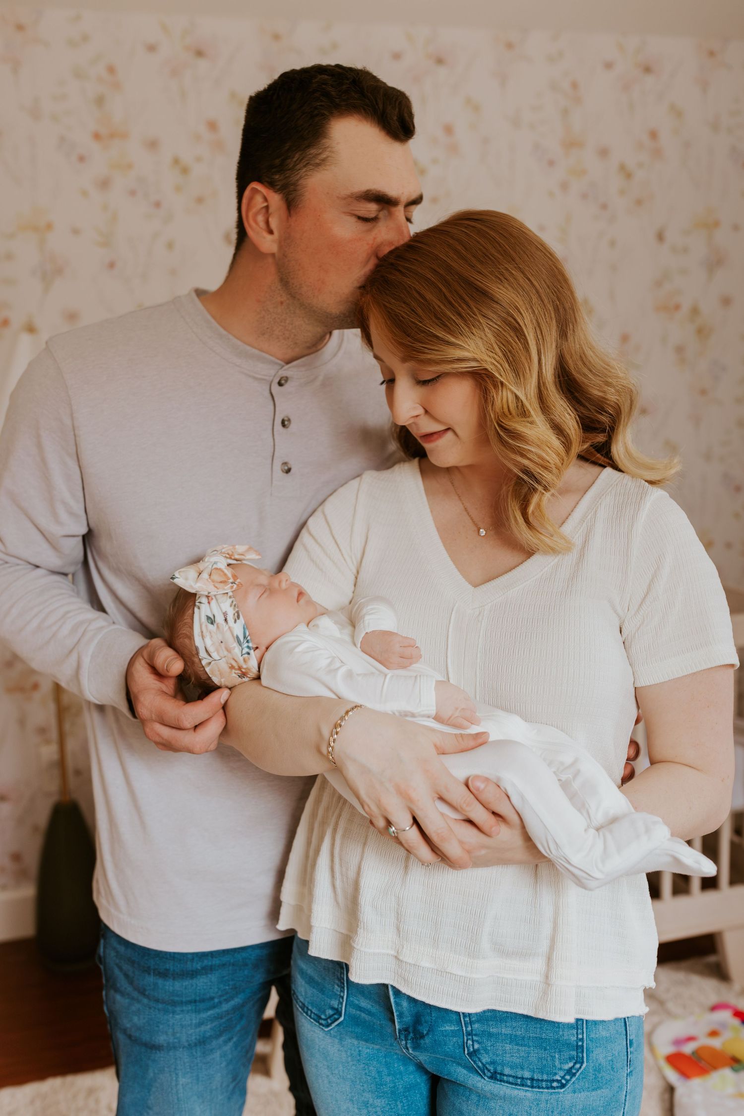 New parents in a floral nursery, father kissing mother's head as she cradles their sleeping newborn baby girl.