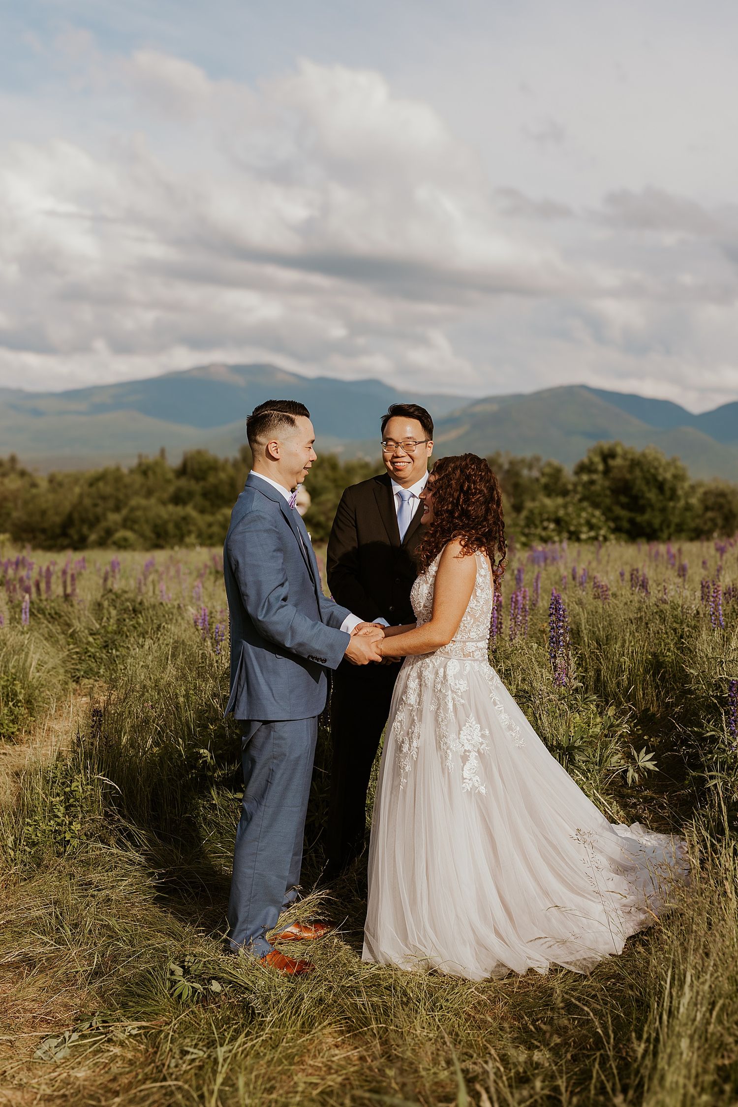 Couple exchanges vows in a lupine field with mountain backdrop, bride in lace gown, groom in blue suit.