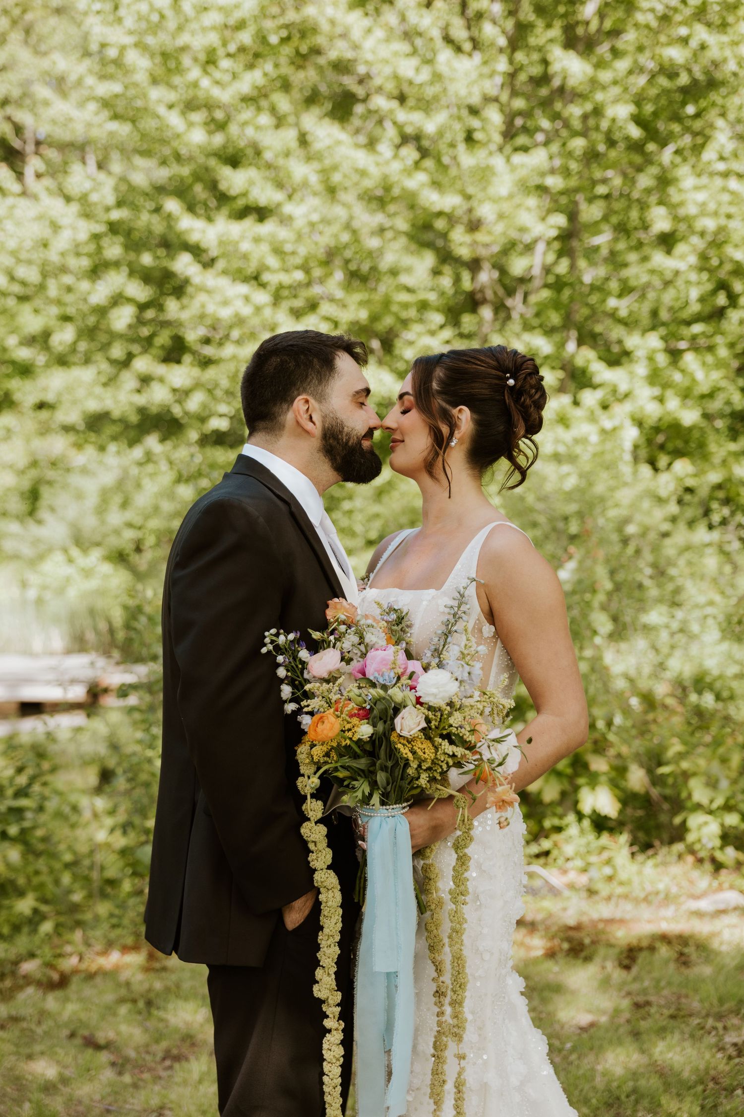 Bride and groom share a romantic moment in a forest setting, bride holding a colorful bouquet with blue ribbons.
