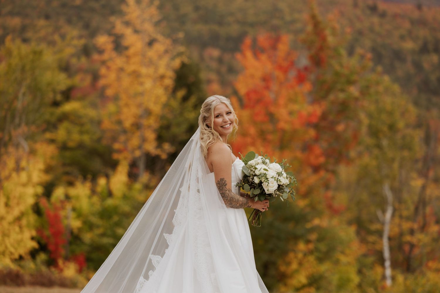 Bride in white gown and veil holds bouquet amid vibrant autumn foliage backdrop.