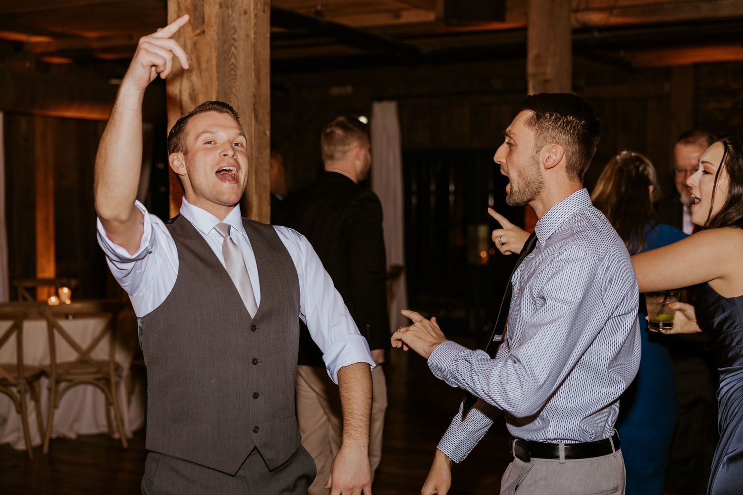 Two men dancing energetically at a wedding reception in a rustic barn venue.