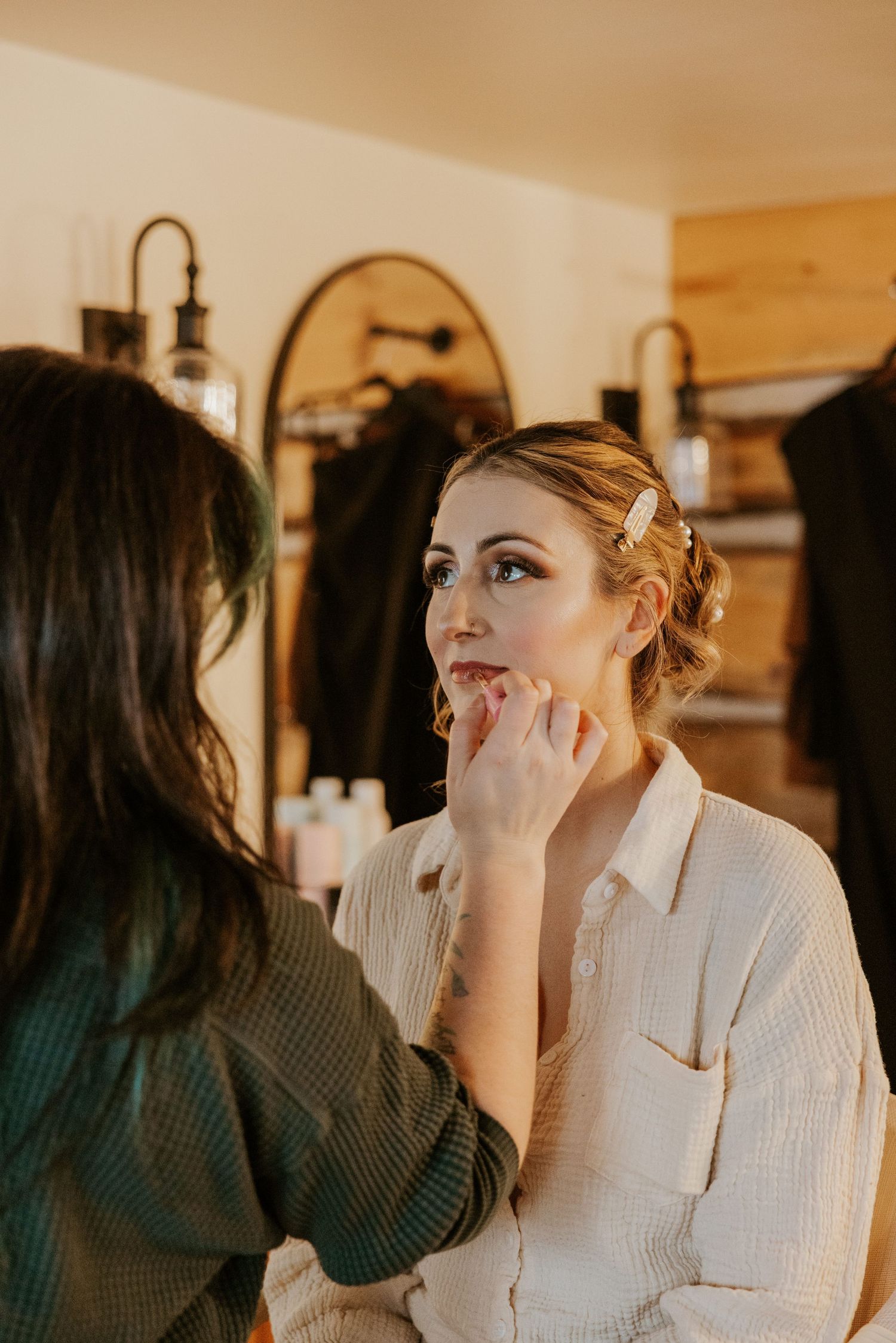 Makeup artist applying finishing touches to bride's face in warm rustic dressing room.