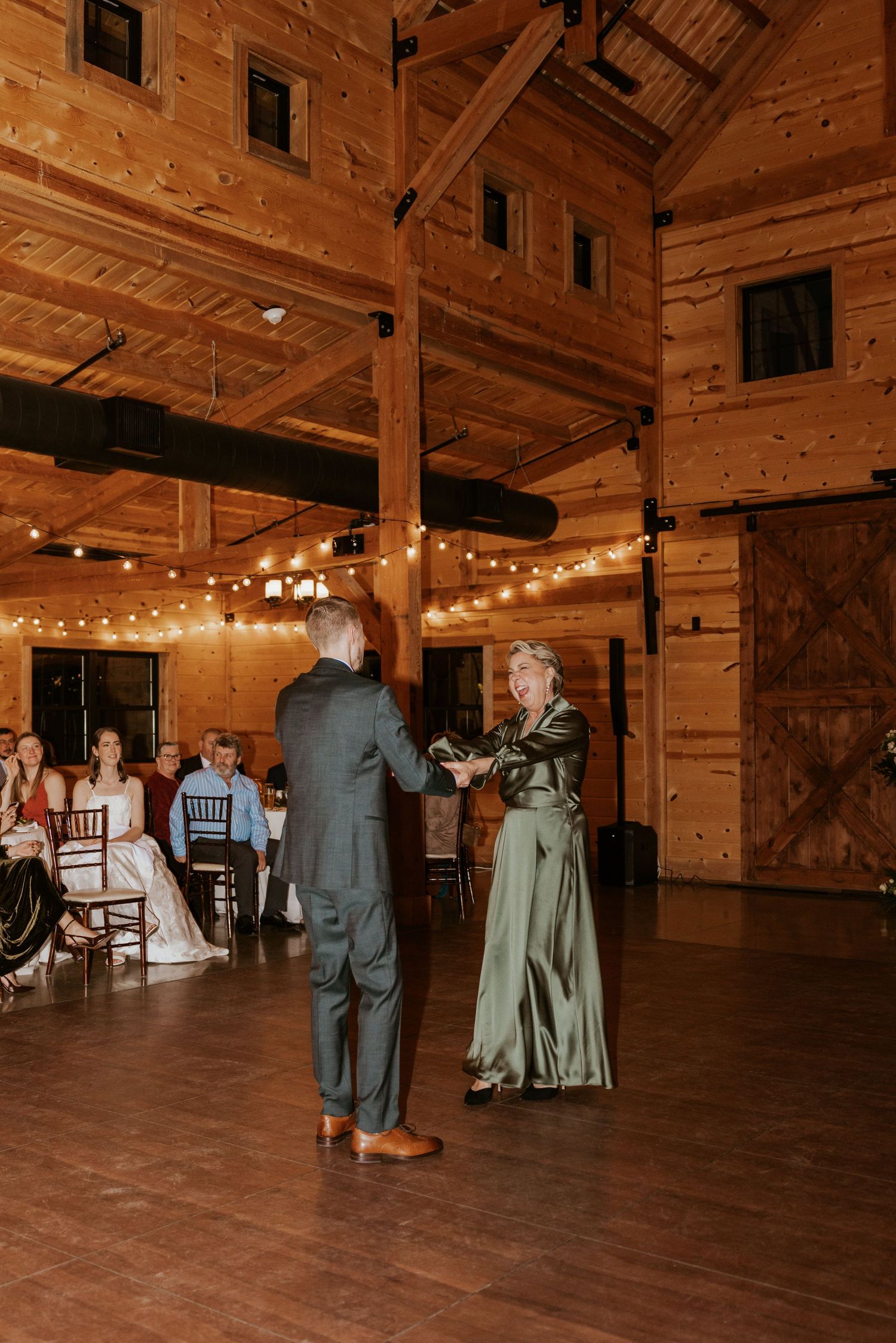 Wedding guests in formal attire dance on a rustic wooden dance floor illuminated by string lights in a barn venue.