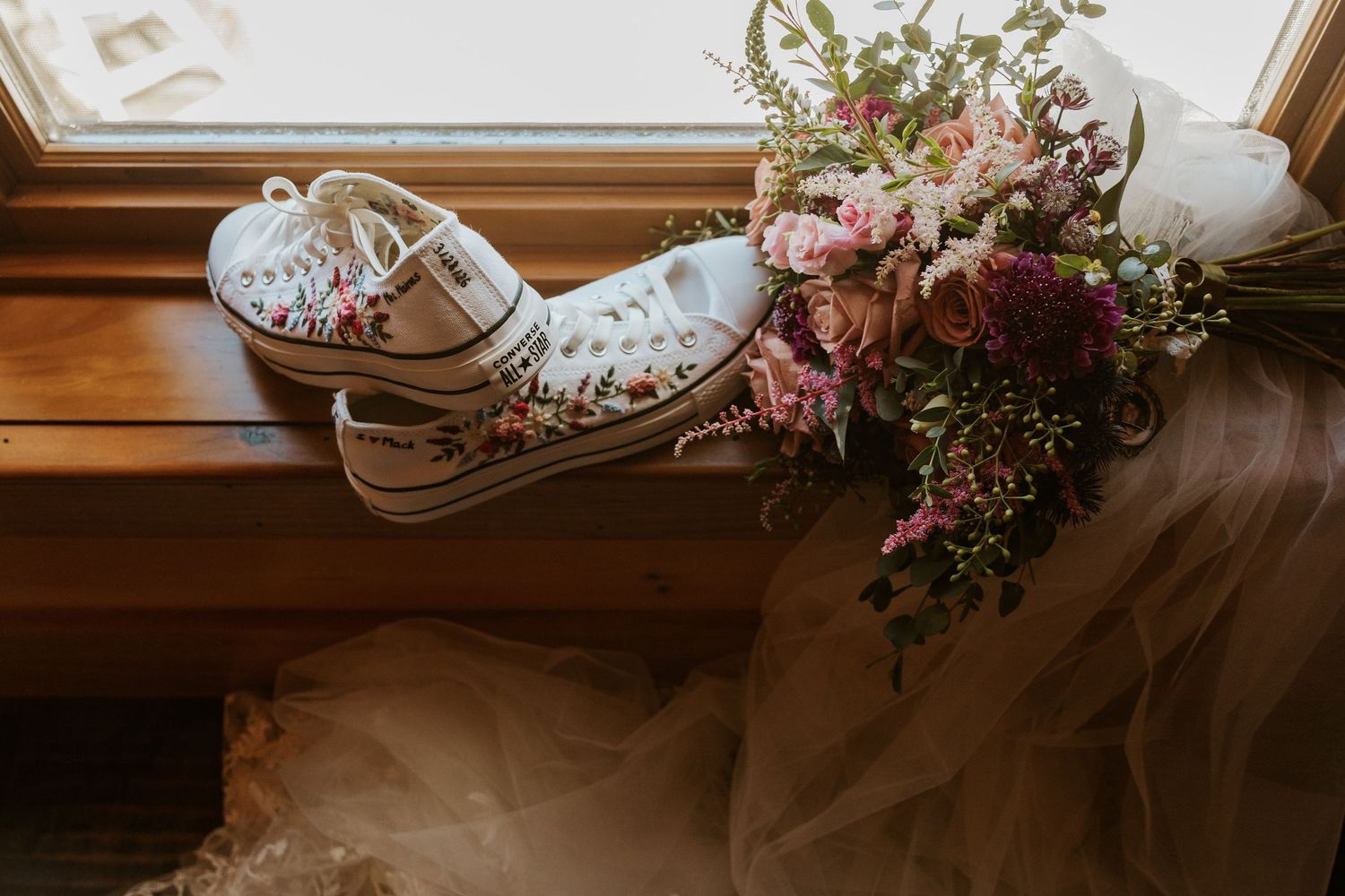 White wedding sneakers adorned with delicate pink flowers and greenery sit on a wooden window ledge.