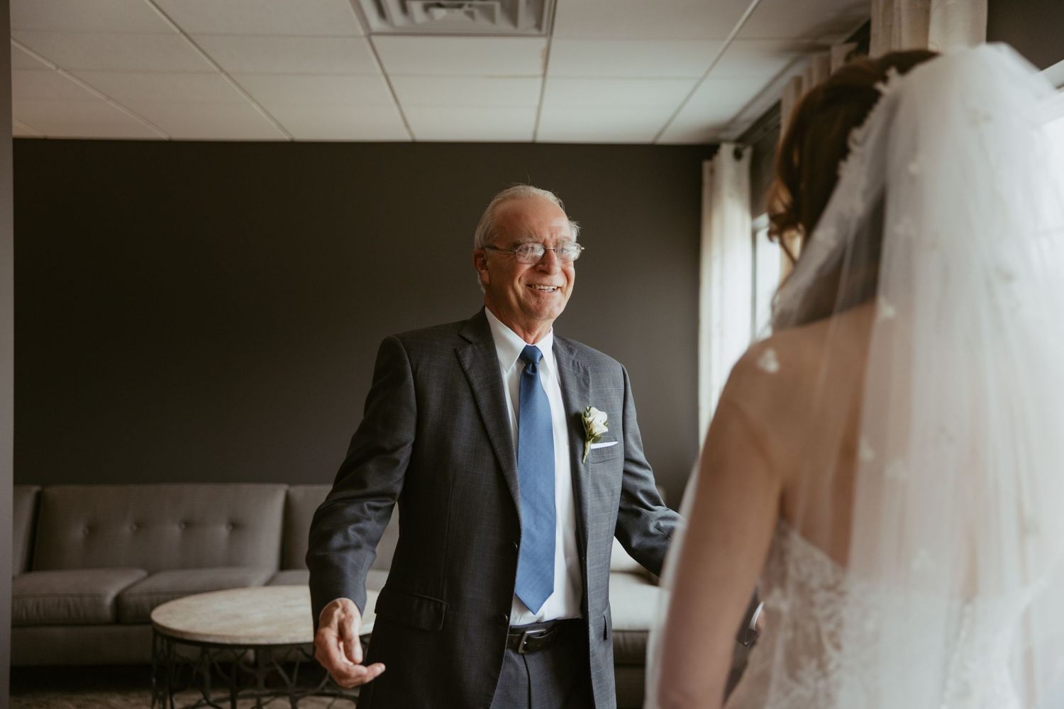A bride in a white wedding dress shares an emotional first look moment with her father in a hotel room.