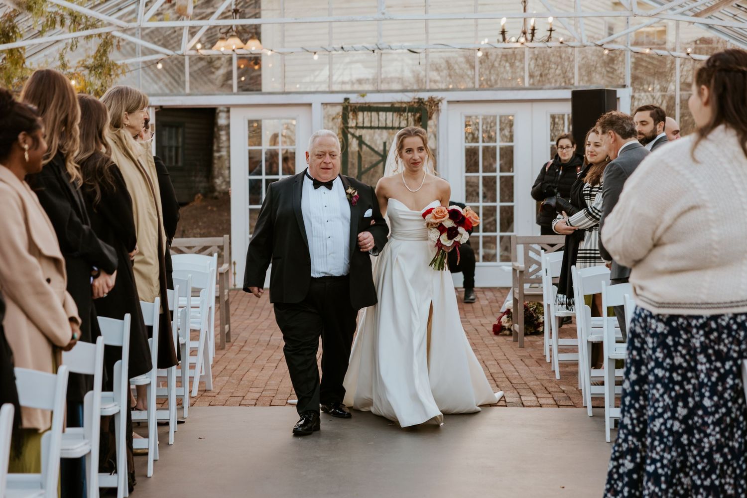 Bride in white satin gown walks down the aisle with her father in a glass greenhouse ceremony venue.