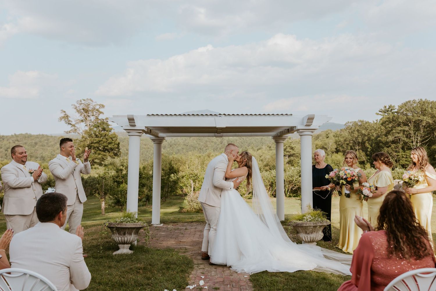 Bride and groom sharing first kiss at outdoor wedding altar with pergola and scenic mountain views behind them.