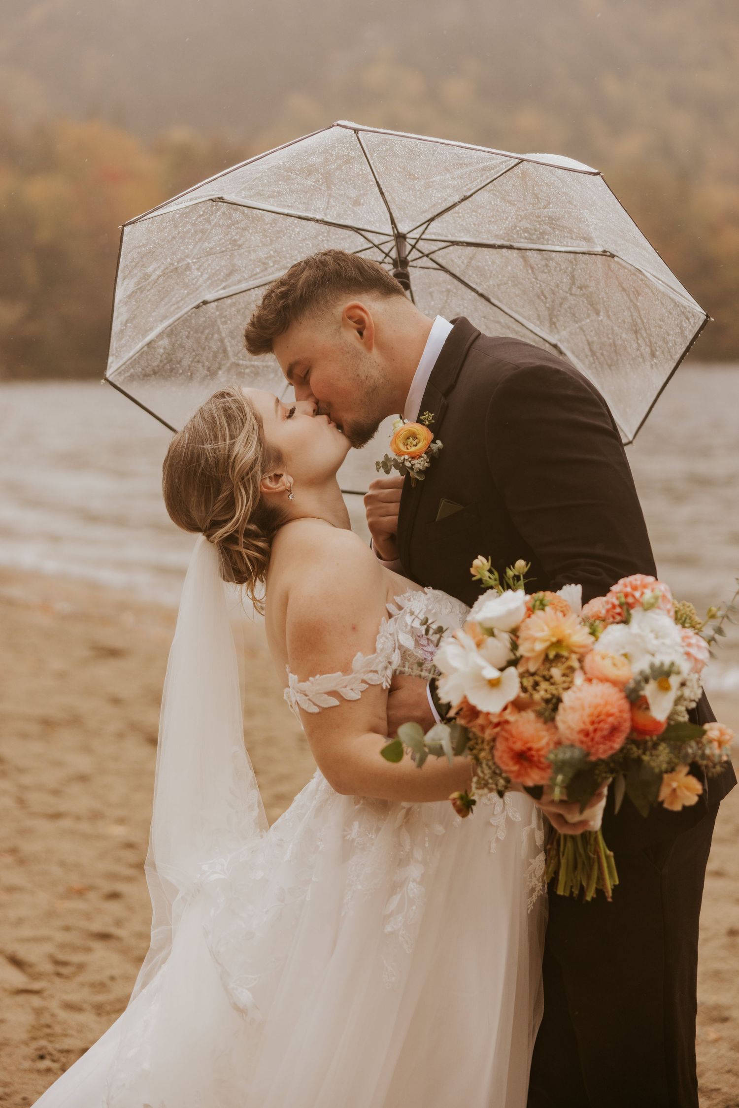 Bride and groom share a kiss under a lace umbrella on a sandy beach with autumn foliage behind them.