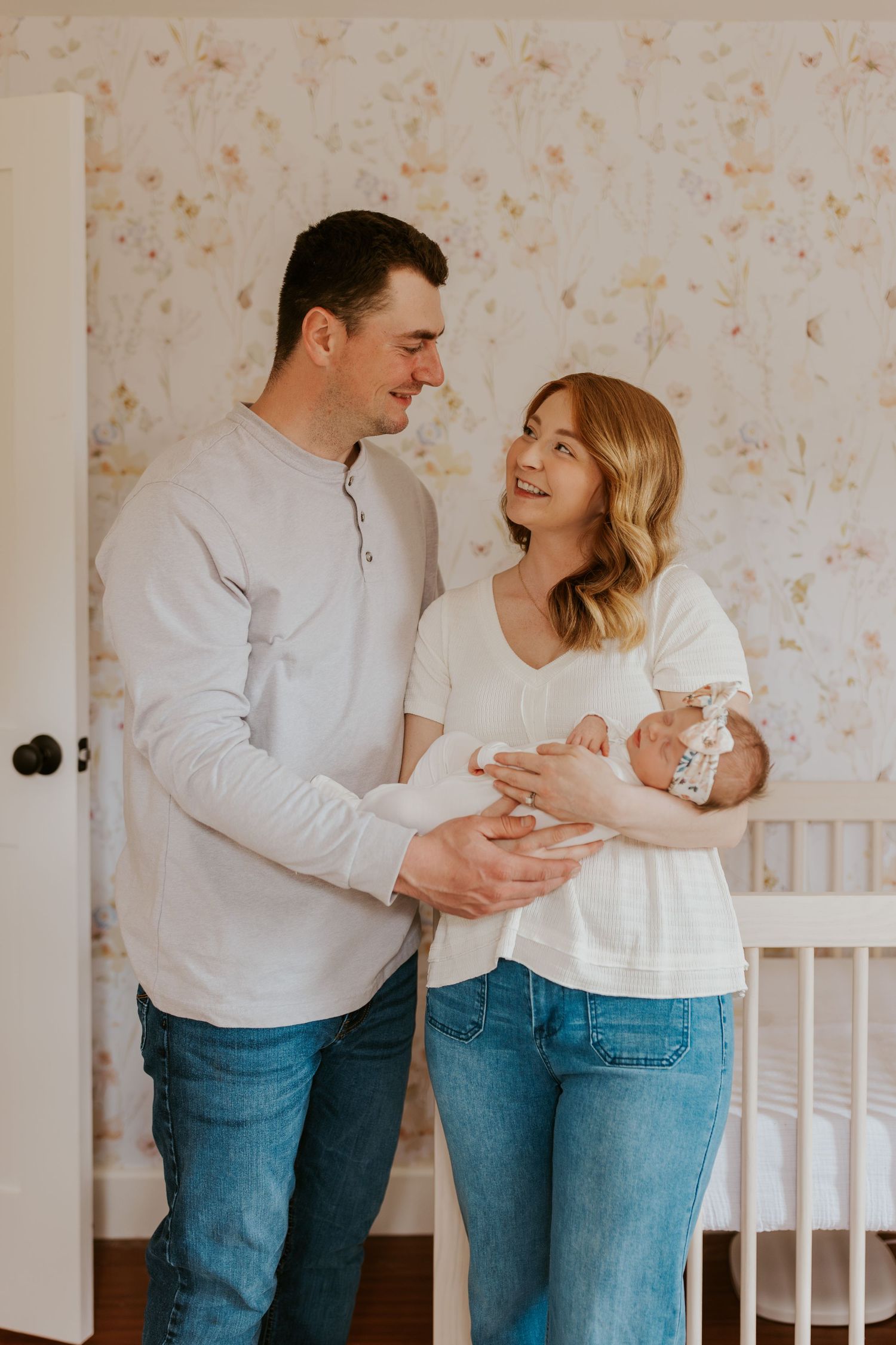 Happy couple standing in a nursery with floral wallpaper, lovingly holding their newborn baby girl together.