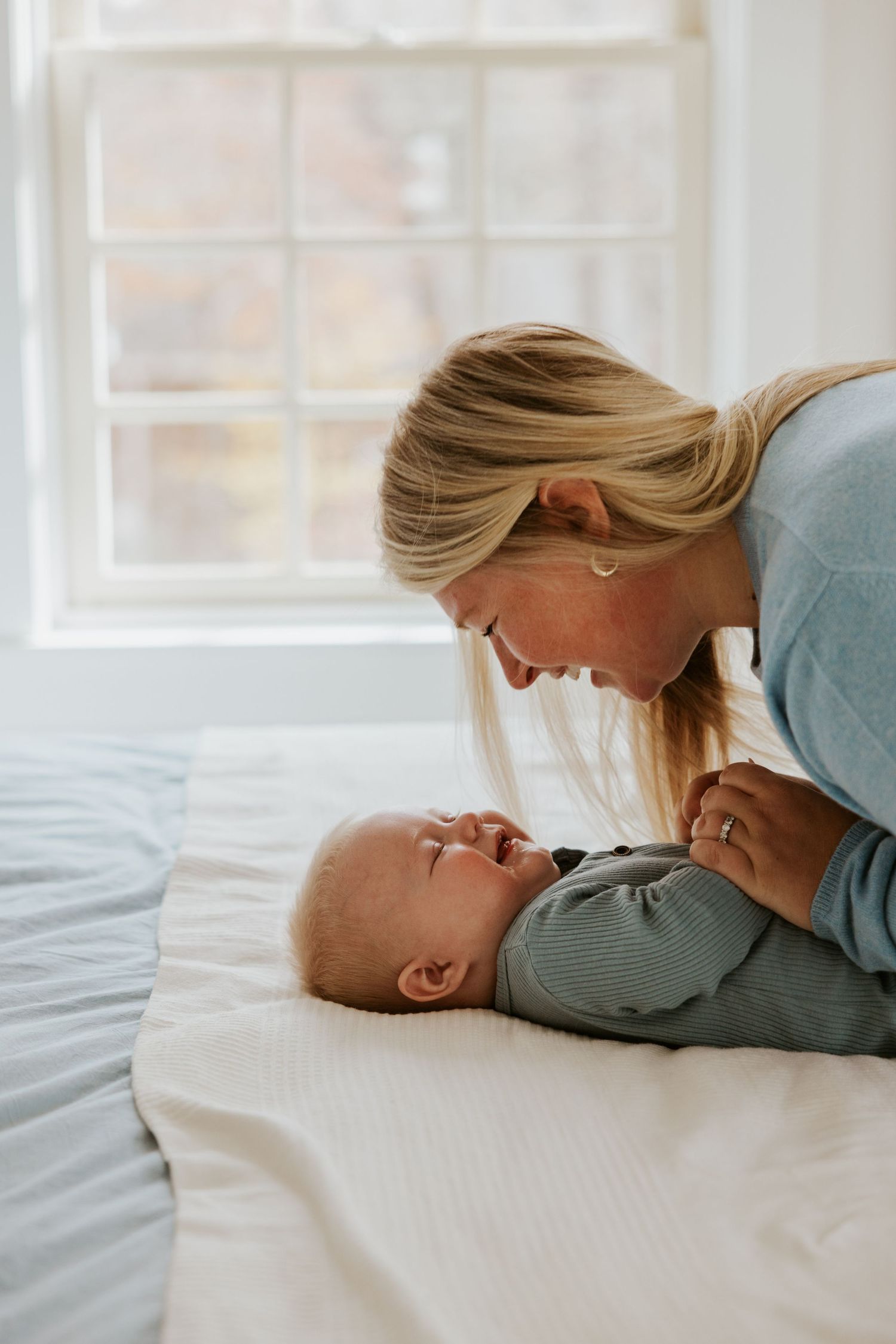 Blonde mother leaning over laughing baby lying on white bed near bright window.