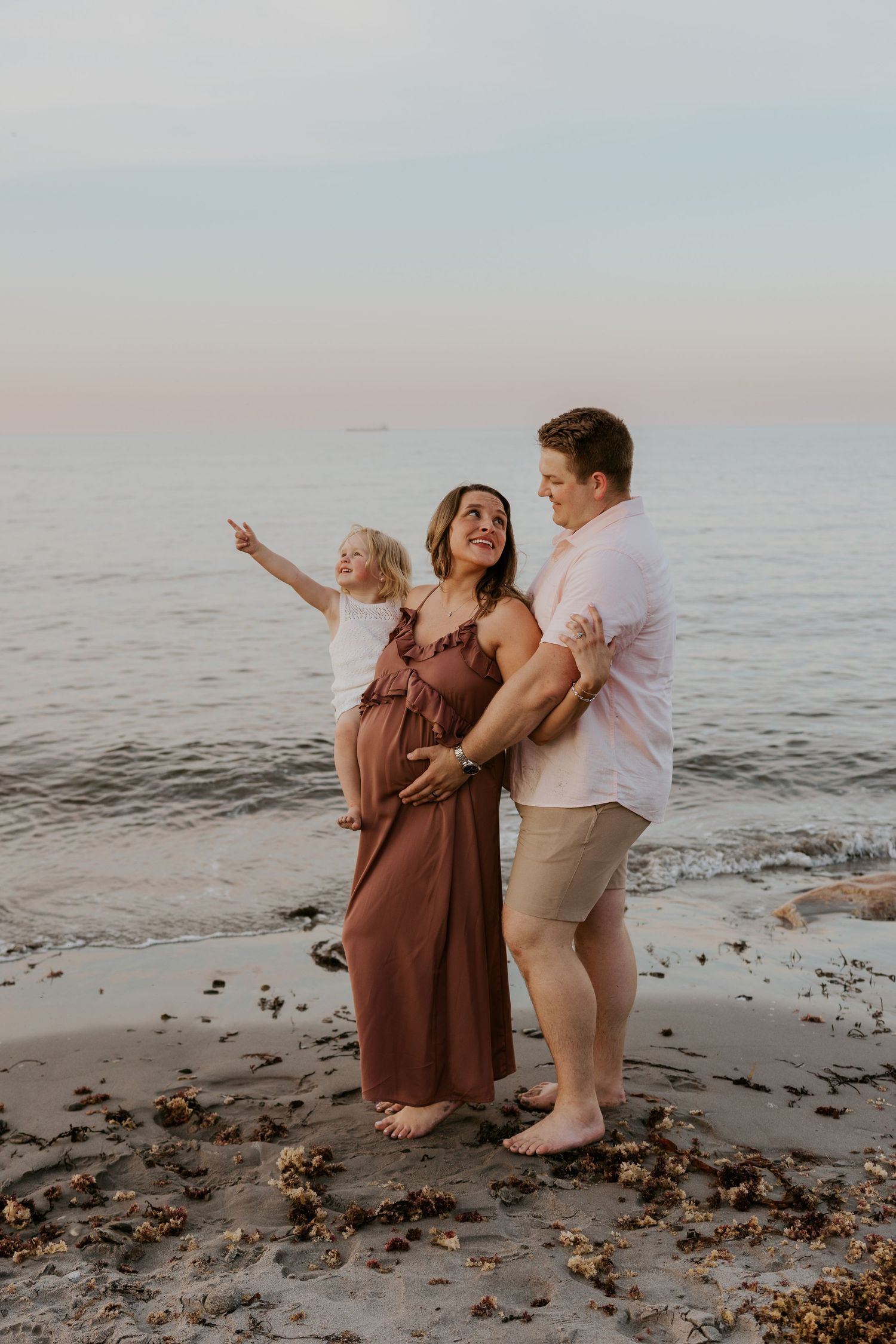 Pregnant woman in brown dress with husband and young child pointing at ocean at sunset.