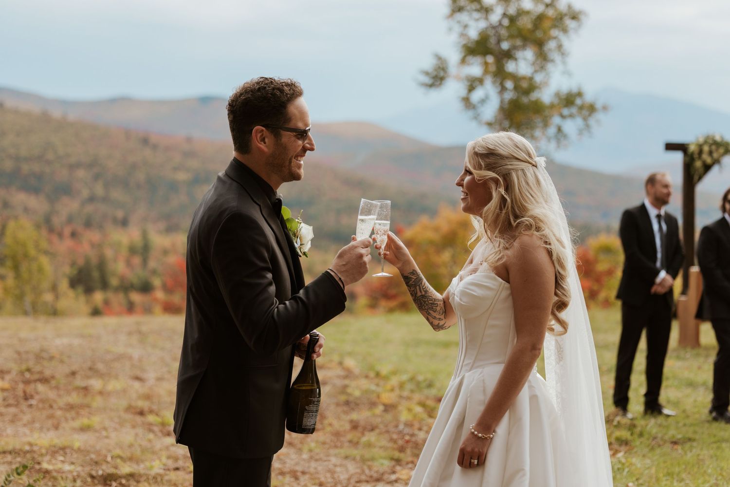 Bride and groom toasting with champagne glasses at outdoor fall wedding with mountain backdrop.