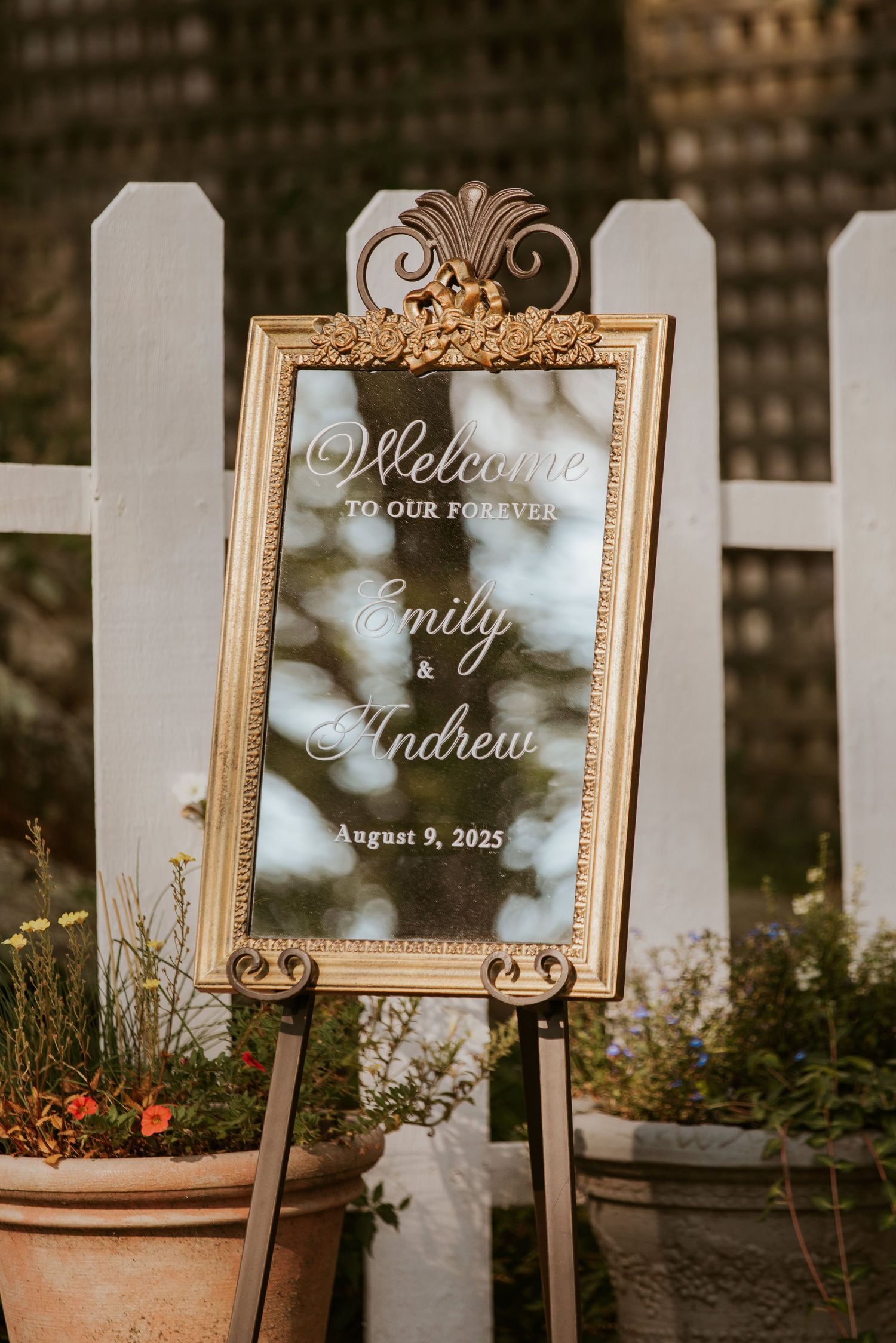 Gold ornate framed wedding welcome mirror sign for Emily and Andrew on August 9 2025 displayed on easel outdoors.