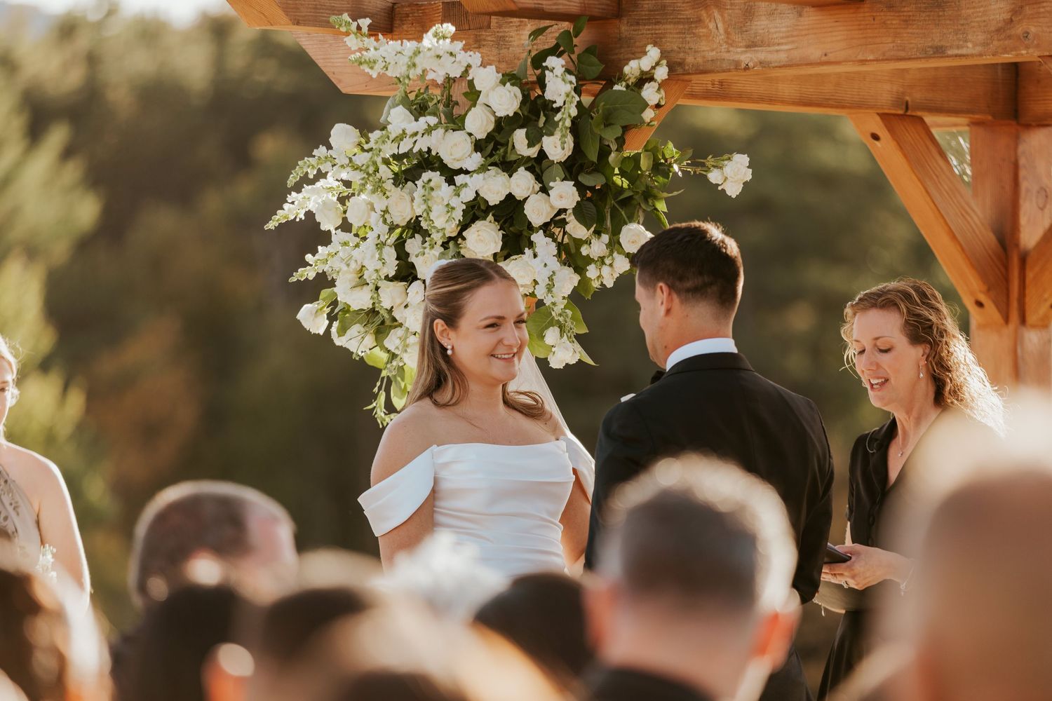 Bride smiling at groom during outdoor ceremony under white floral-decorated wooden pergola arch.