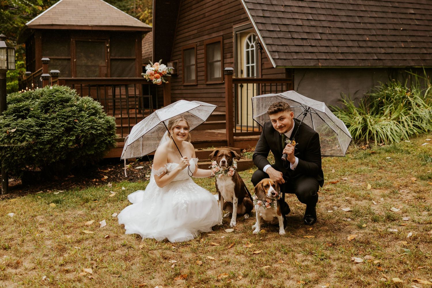 Bride and groom pose with two dogs under white umbrellas outside a rustic cabin during a rainy fall wedding.