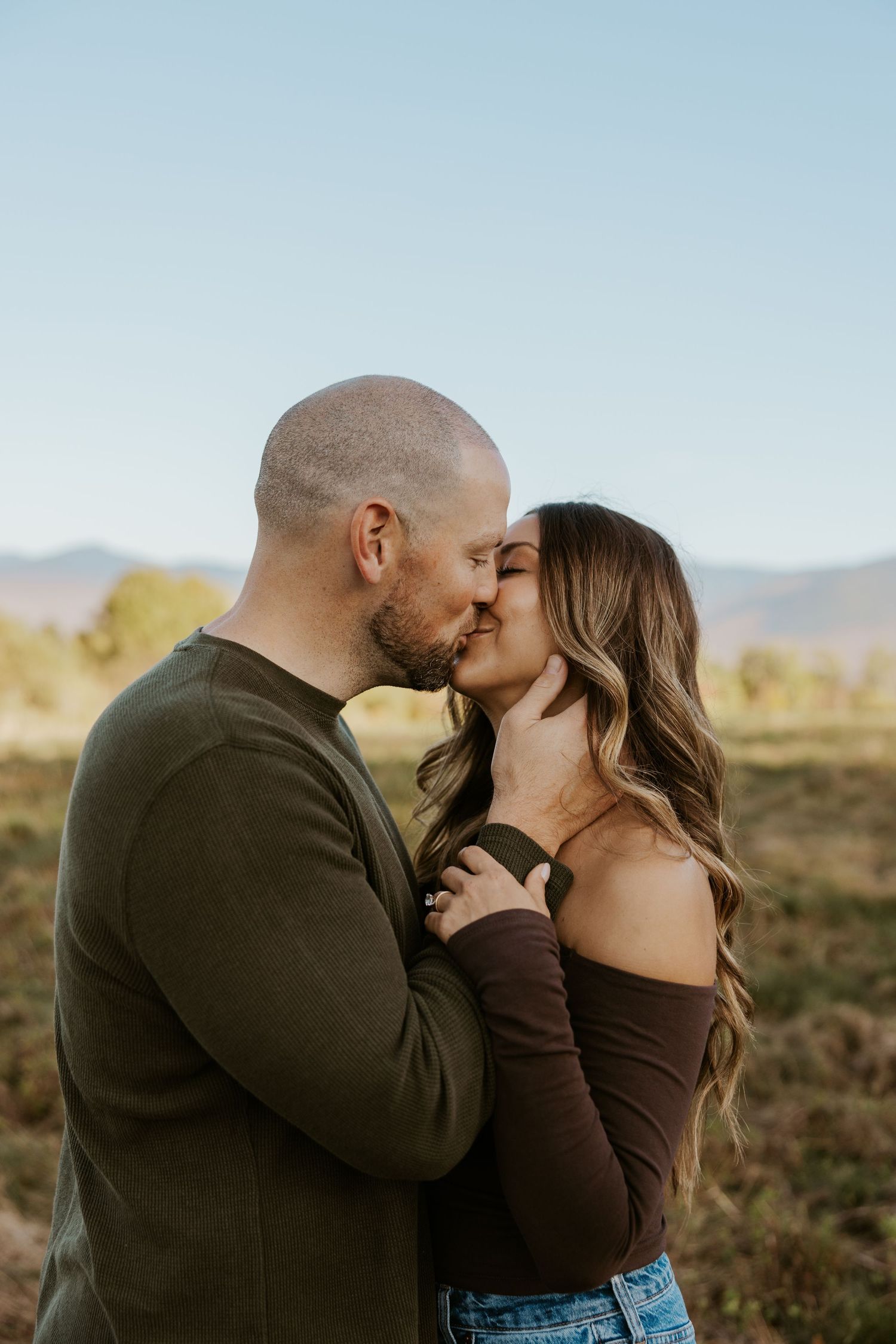 Couple sharing a romantic kiss in an open field with mountains in the background.