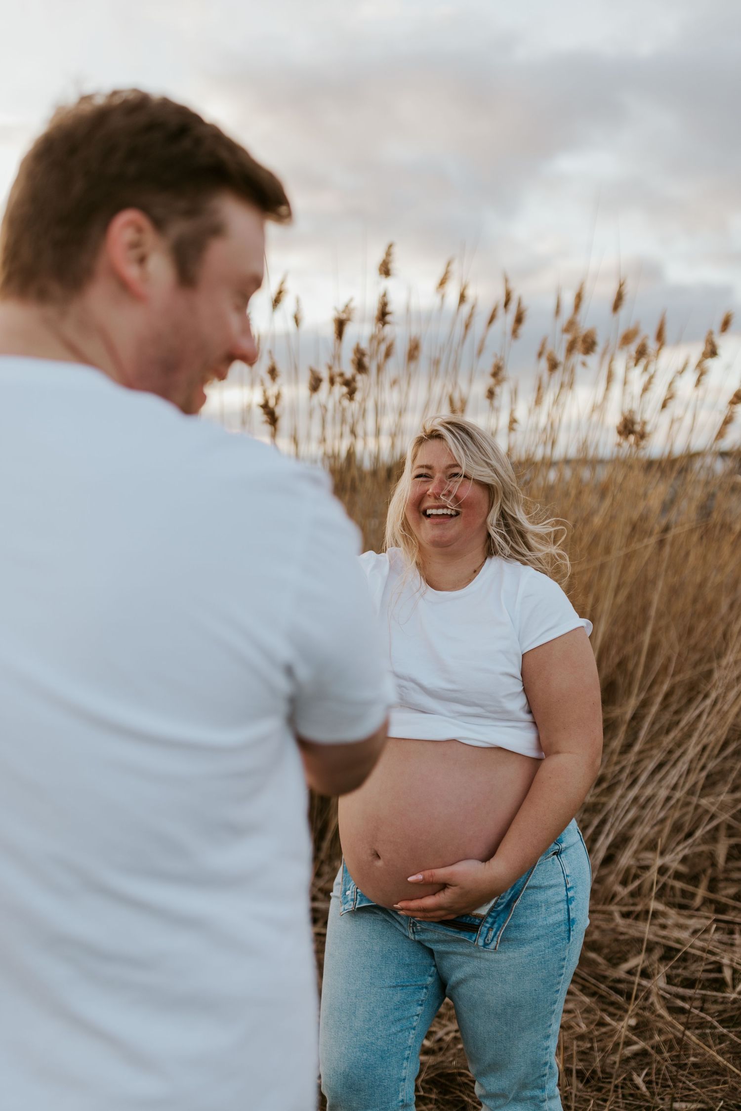 Laughing pregnant woman in white crop top holding belly with partner in field.