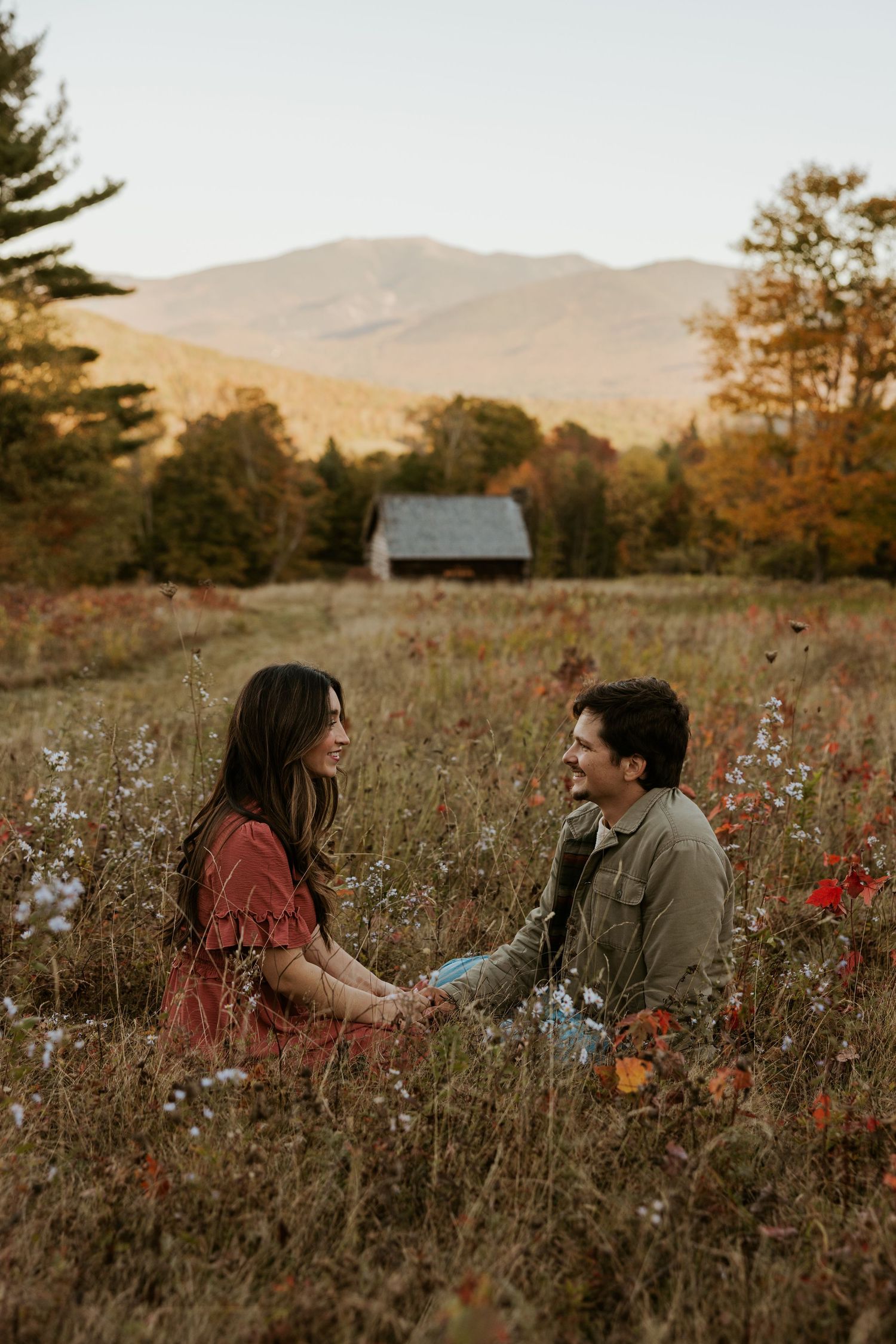 Couple sitting in autumn wildflower field holding hands, smiling at each other with mountain backdrop.