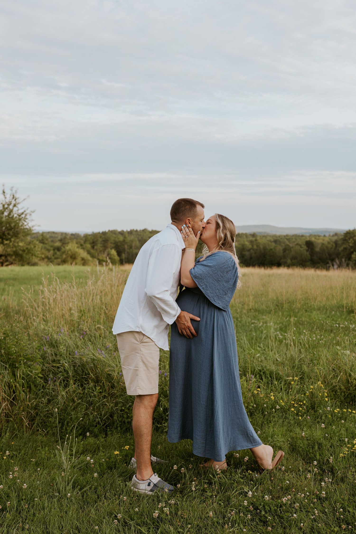 Couple shares a kiss in a green meadow, woman in blue maternity dress, man in white shirt.