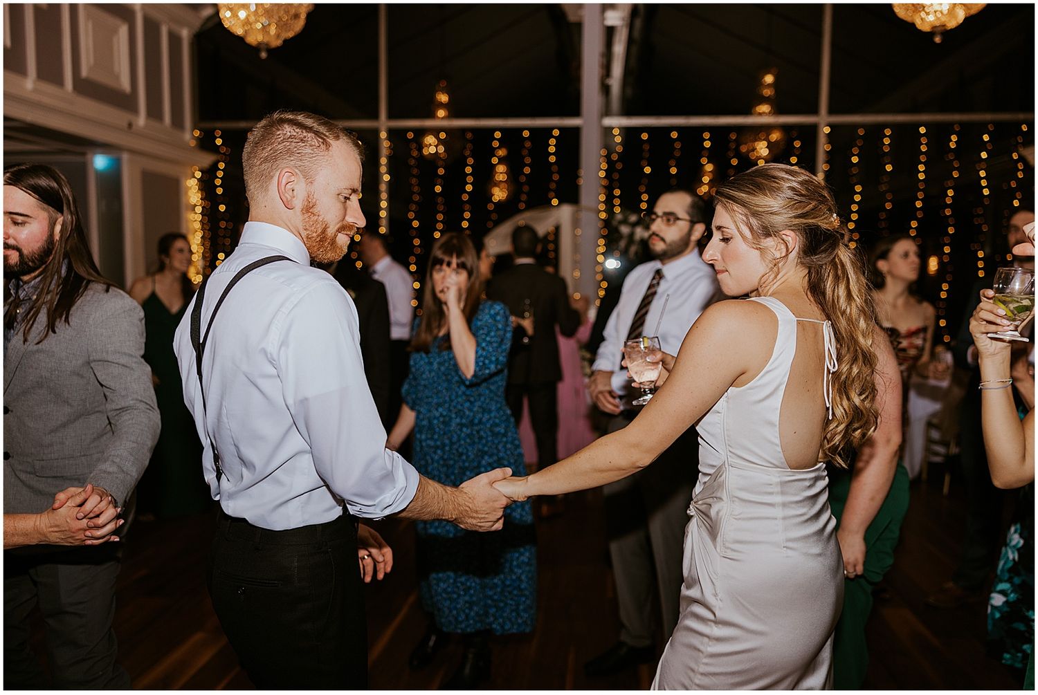 Wedding reception guests dancing under warm string lights and crystal chandeliers.