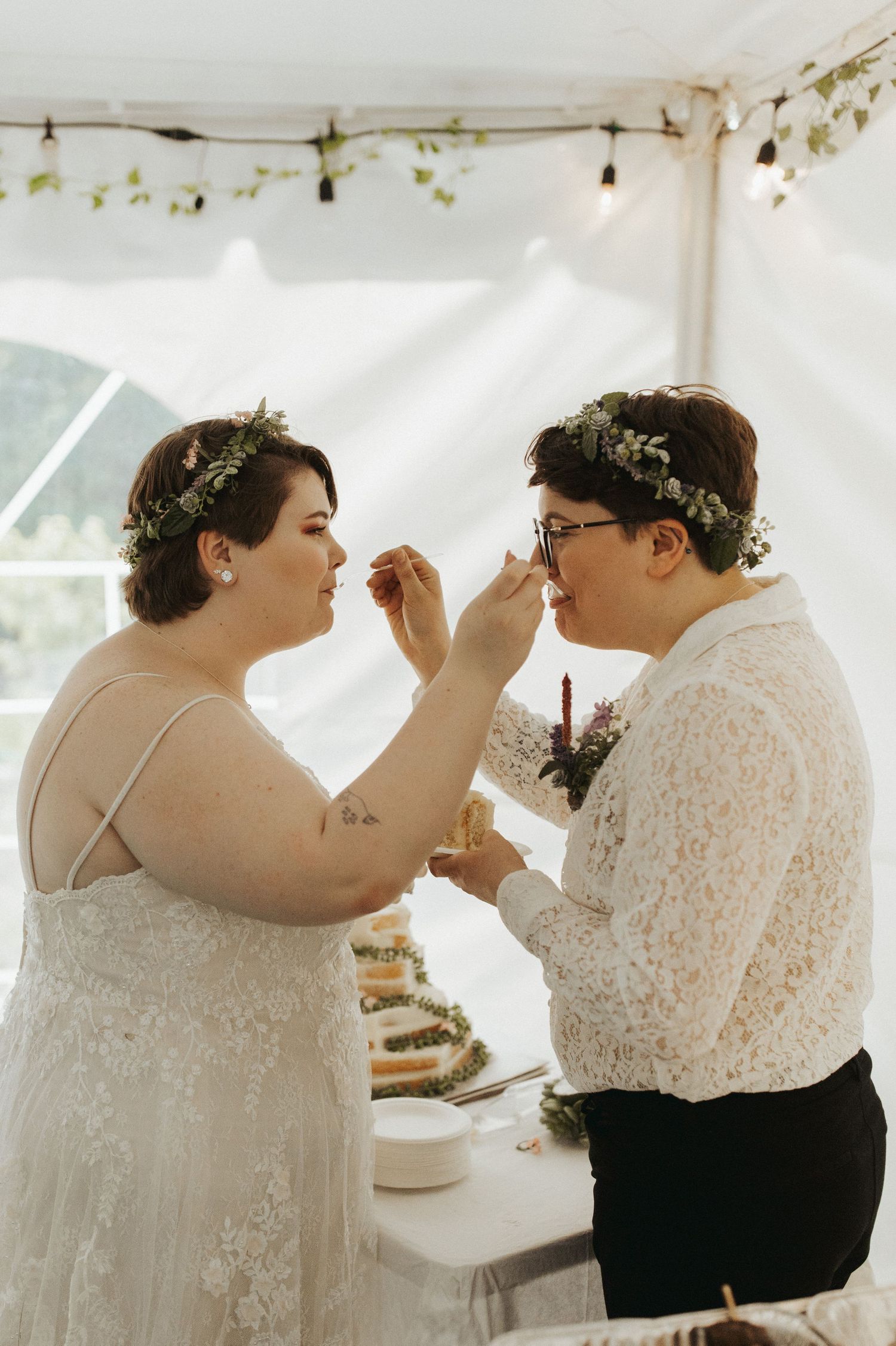 A couple shares cake during their wedding reception under a white tent with string lights.