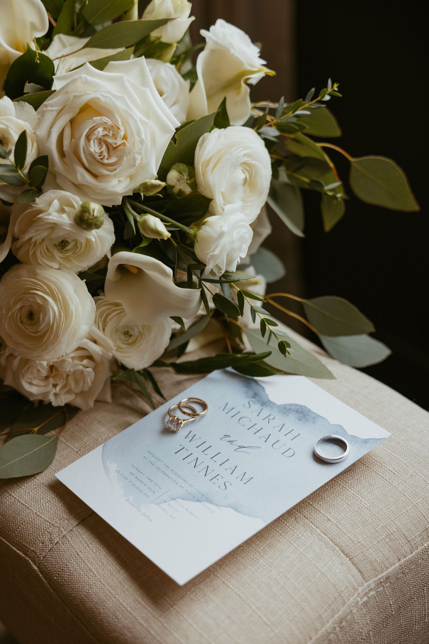 Wedding rings rest on a white card with elegant calligraphy, surrounded by cream roses and eucalyptus on linen fabric.