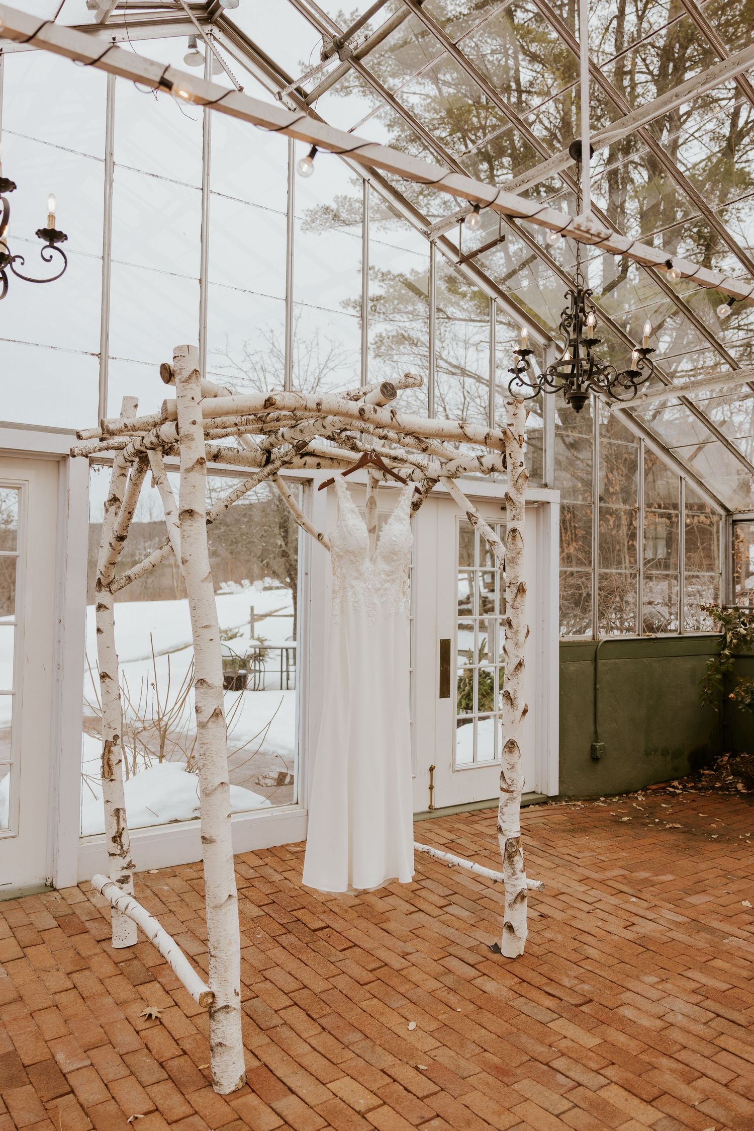 White wedding dress hanging on a rustic birch wood arch inside a glass greenhouse venue with chandeliers.