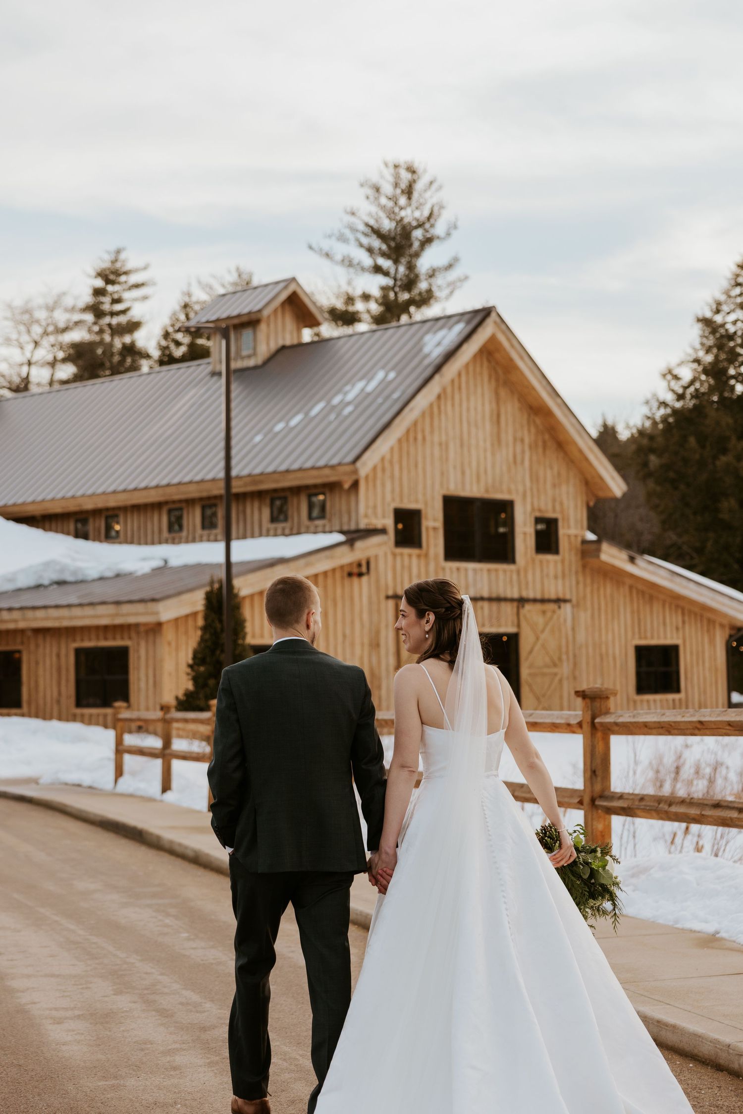 A couple in wedding attire walking towards a rustic wooden barn venue on a snowy winter day.