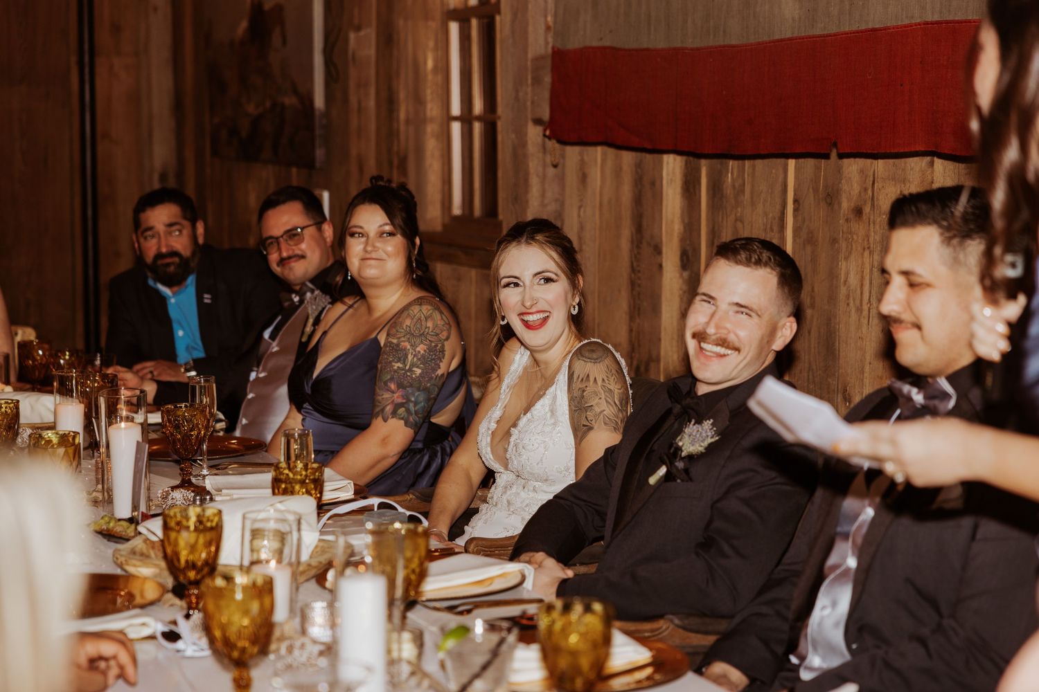 Wedding guests and couple laughing joyfully at a rustic reception dinner table with amber glassware.
