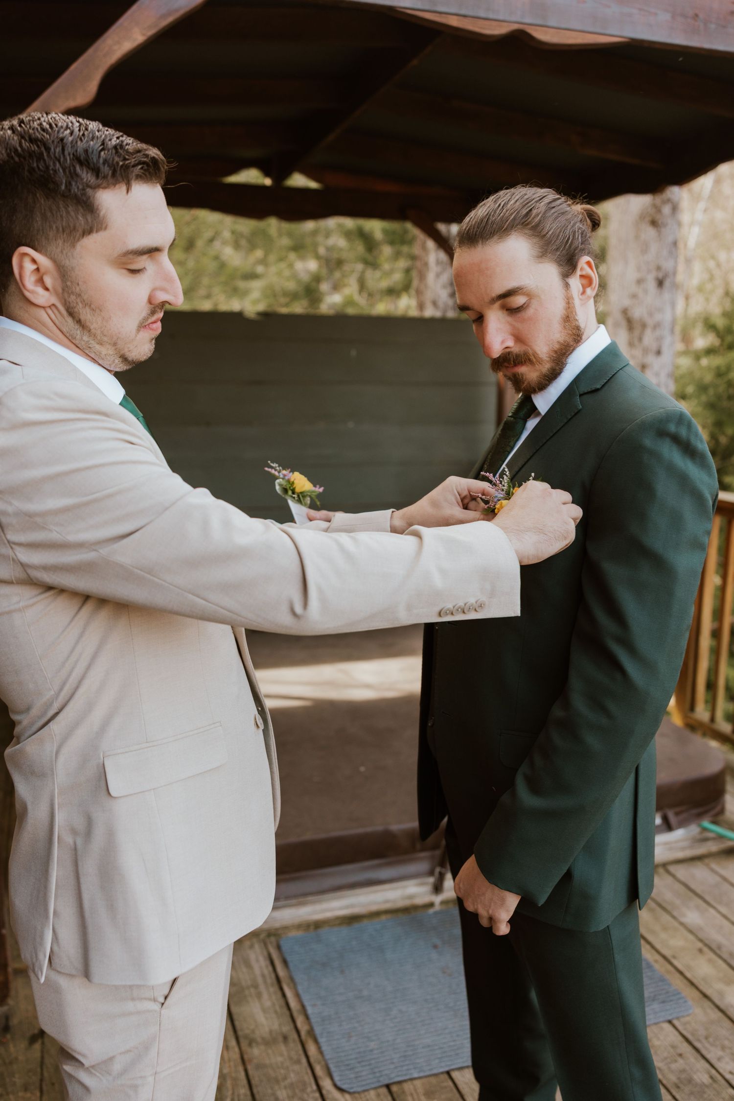 Groomsman in beige suit pins boutonniere on groom in dark green suit at outdoor wedding venue.