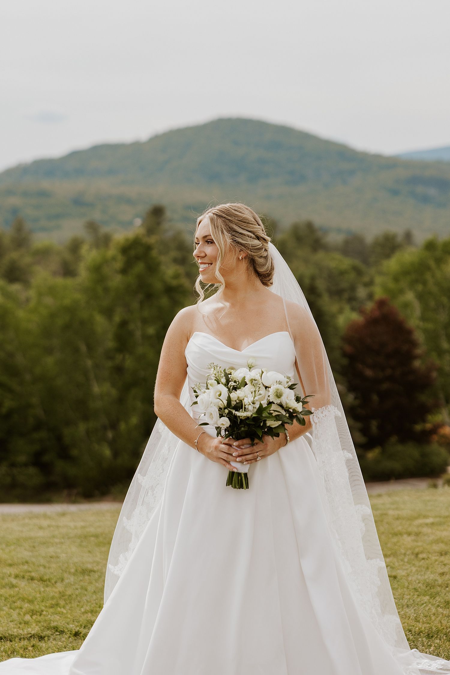 Bride in white ballgown holding white bouquet smiles outdoors with mountain backdrop on her wedding day.