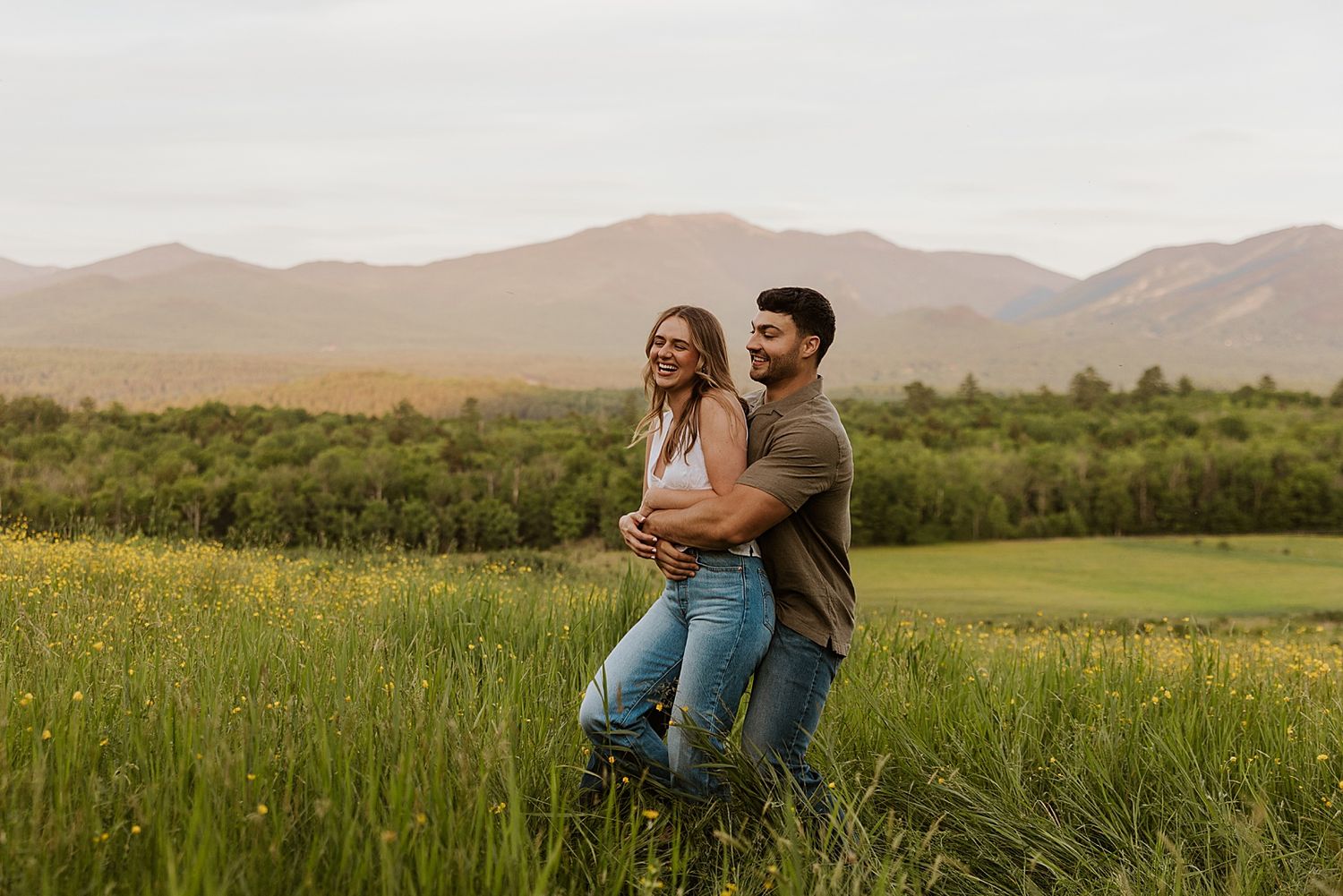 Couple embracing and laughing in a green meadow with mountain views in the background.