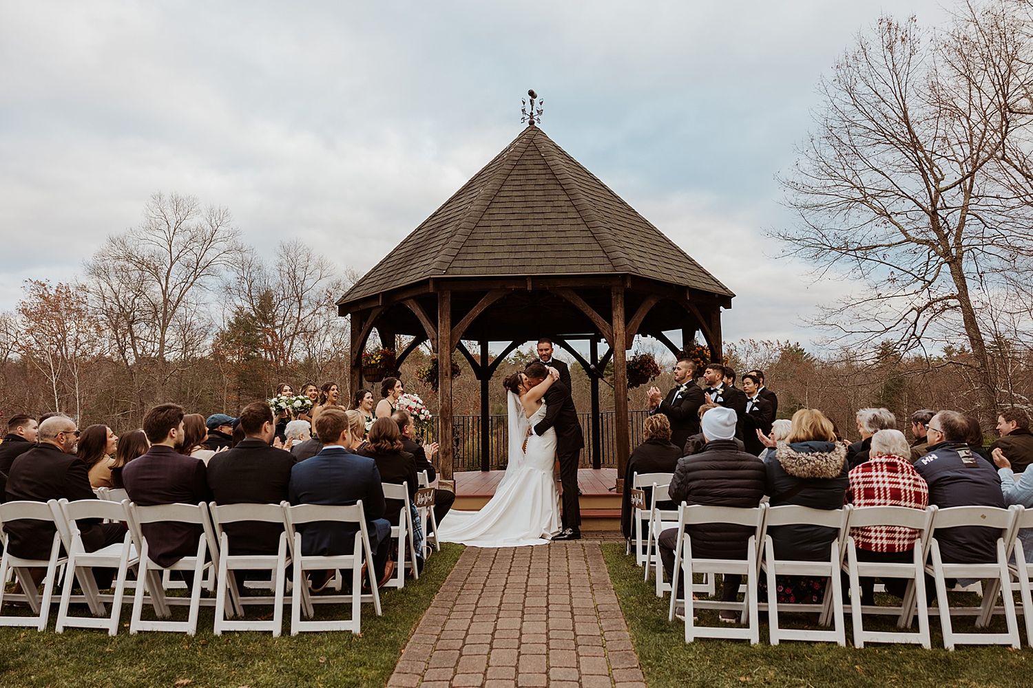 Outdoor NH wedding ceremony at a wooden gazebo with guests seated in white chairs on a brick path surrounded by autumn trees.