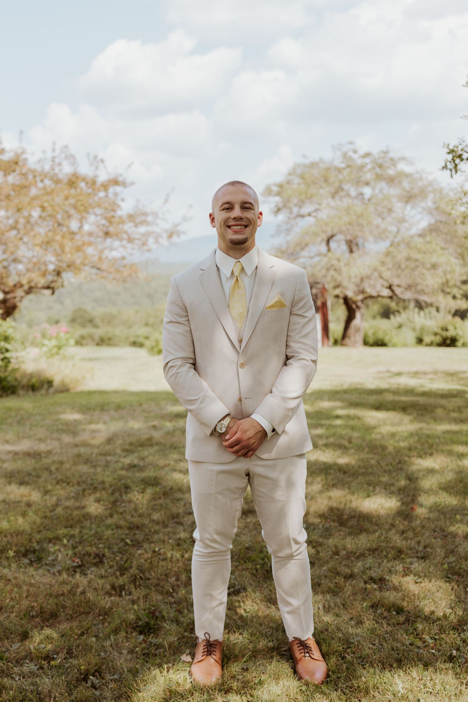 Dapper groom in beige suit with yellow tie and brown shoes smiles confidently on wedding day.