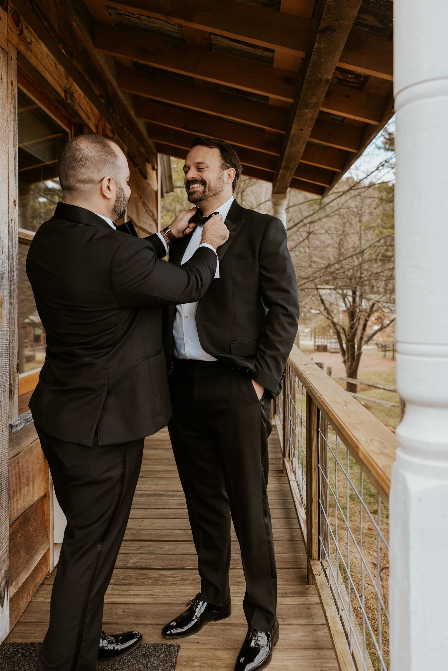 Two groomsmen in black tuxedos share a joyful moment as one adjusts the others bow tie on a wooden porch.