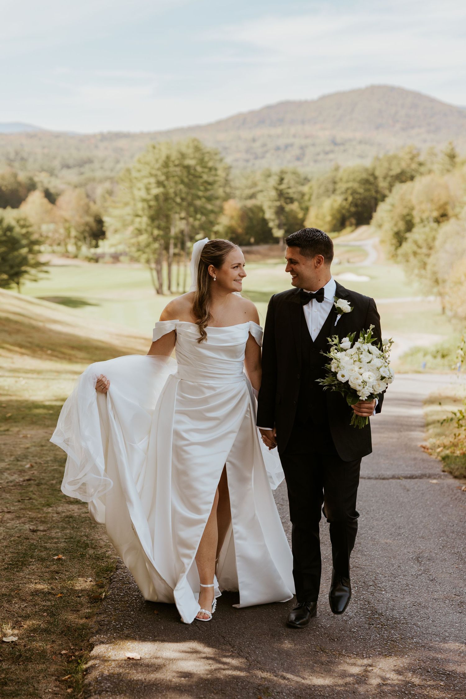 Bride and groom walking hand-in-hand on path with mountain and golf course backdrop on wedding day.